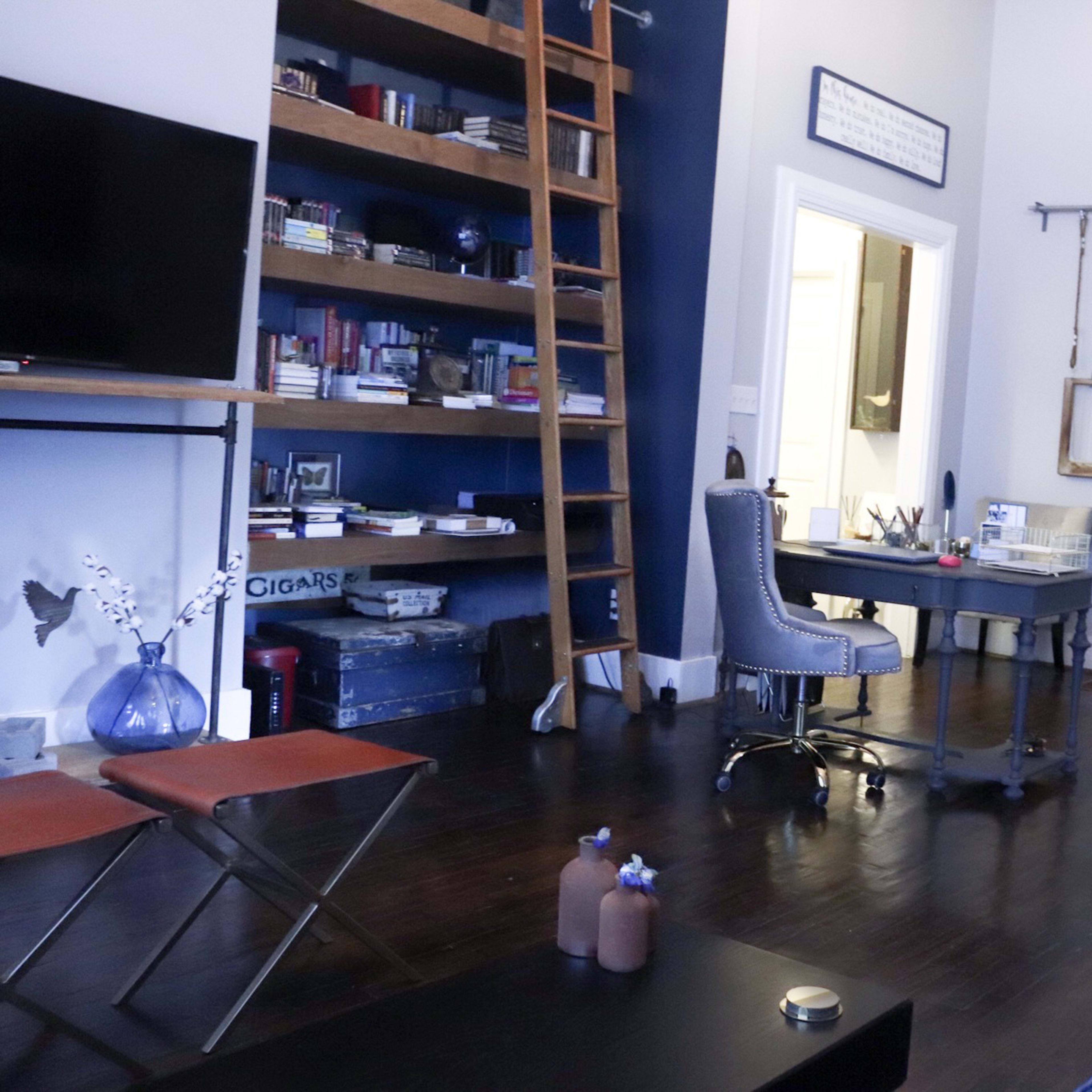 A well-furnished living space featuring a blue couch, a wooden ladder against a bookshelf, a desk with a chair, and a door leading to another room.