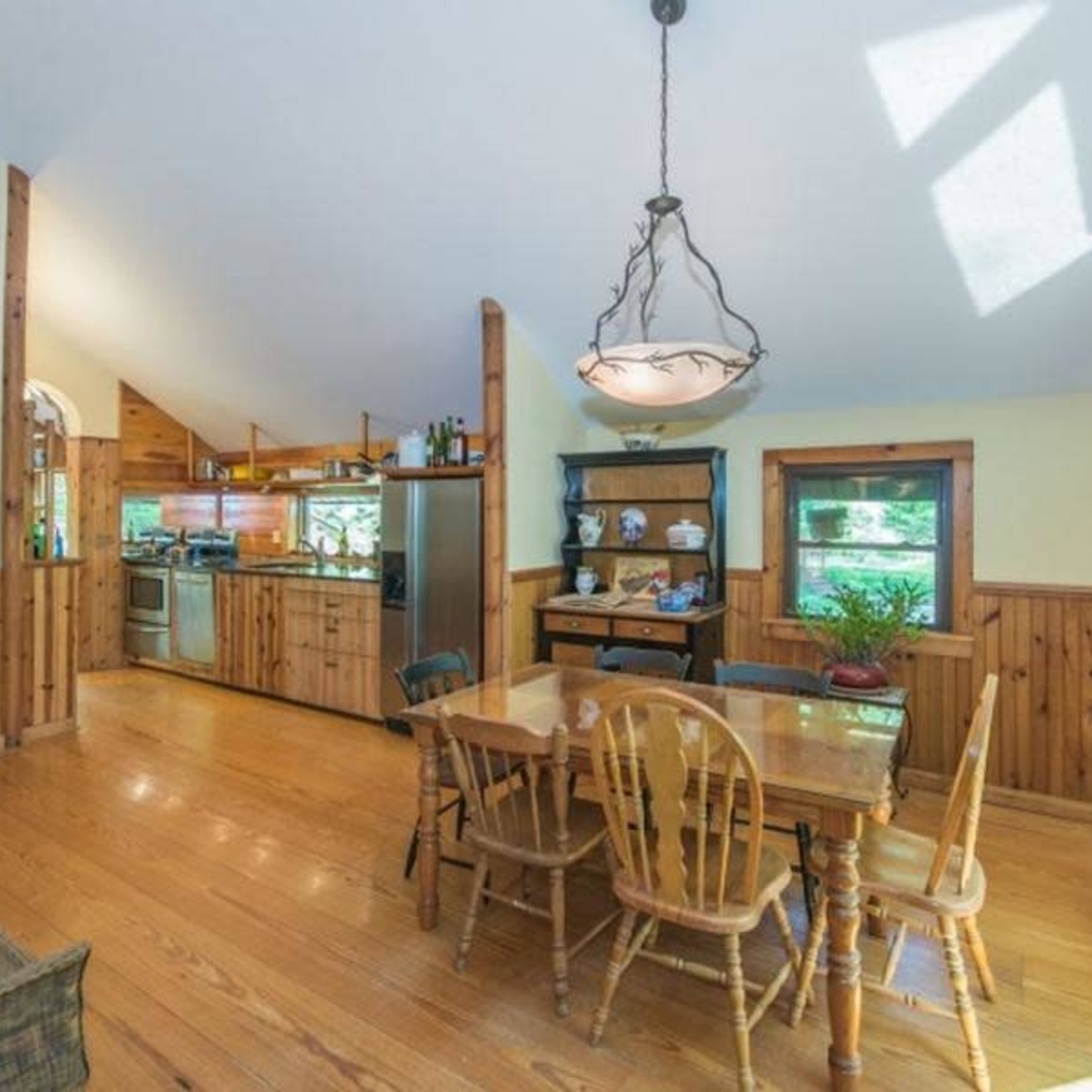 A bright kitchen and dining area featuring wooden floors, a table with four chairs, and a window overlooking greenery.