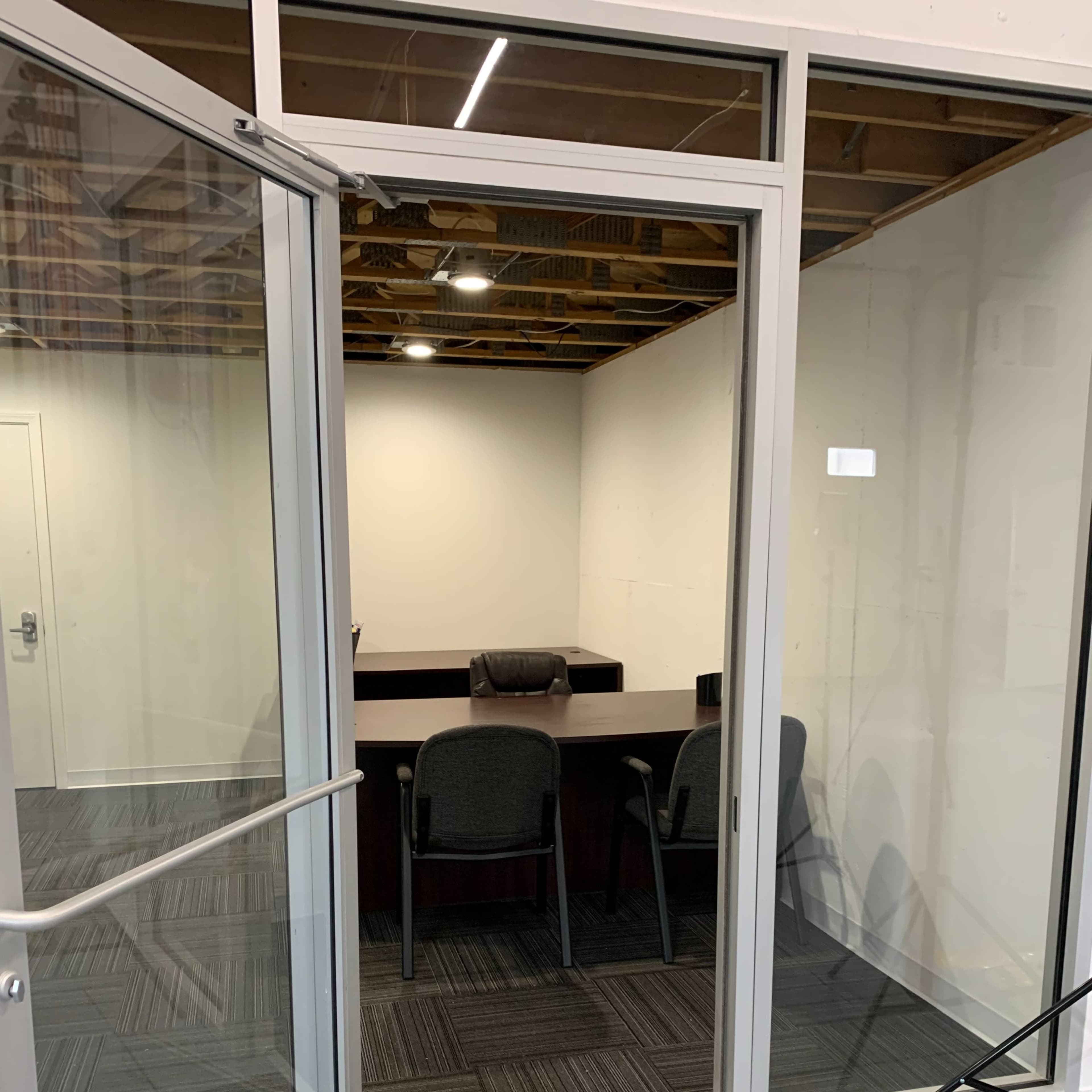 An office space features a large wooden table with two chairs, visible through a glass door, with a gray carpet and exposed overhead beams.
