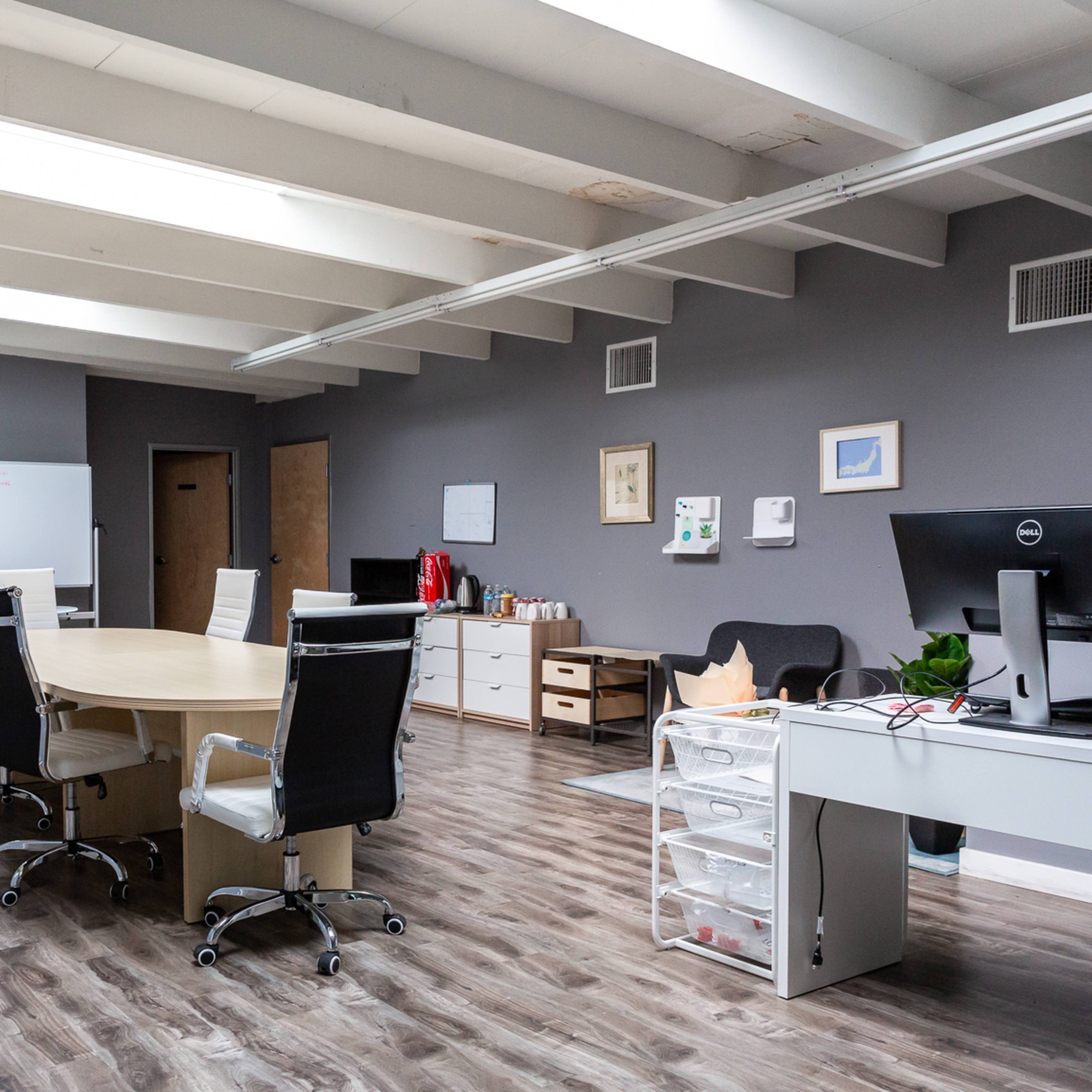 A modern office space with a large round table, several chairs, and a desk with a computer, featuring light wood flooring and gray walls.