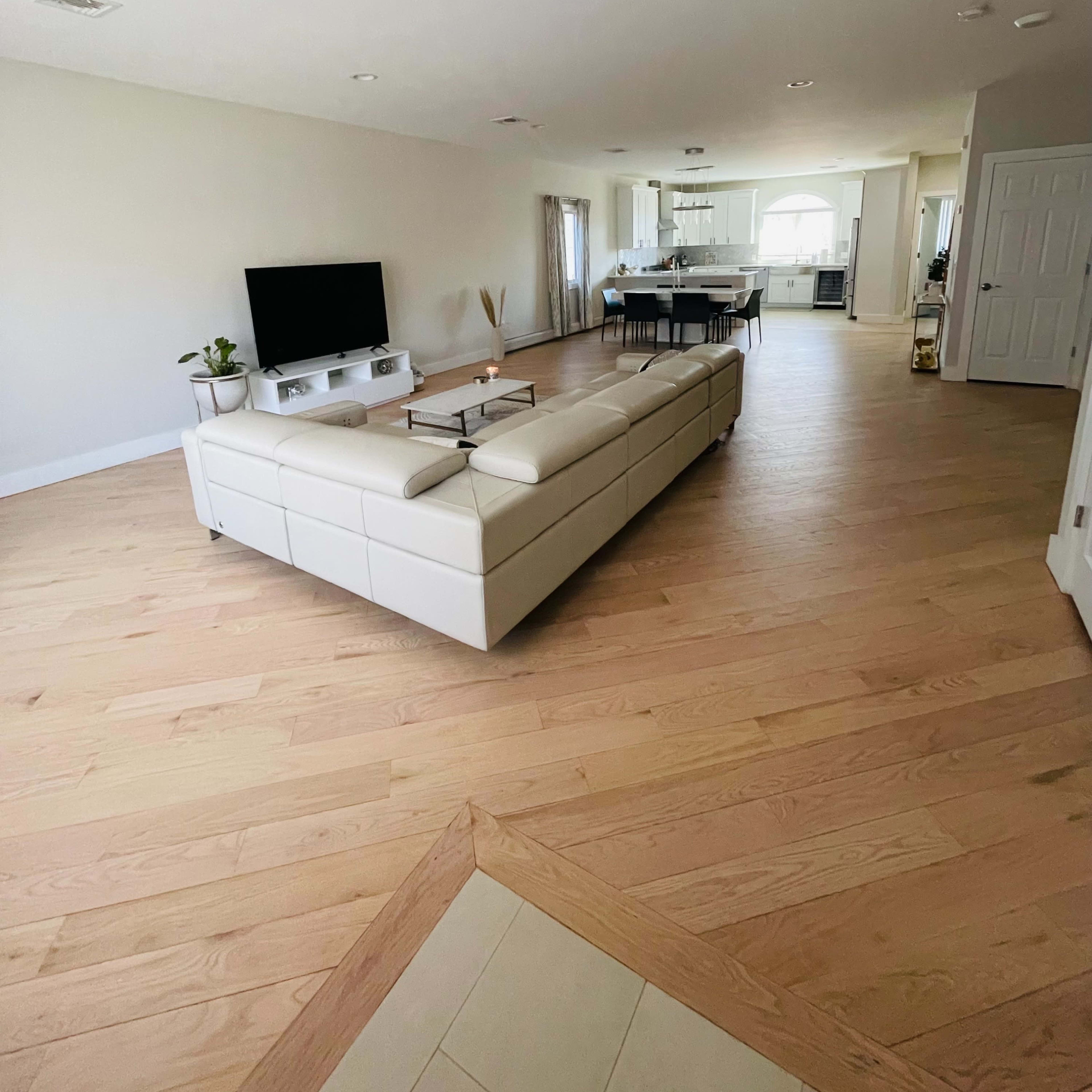 A spacious, modern living room features a white sectional sofa and a television, overlooking an open kitchen area with large windows.