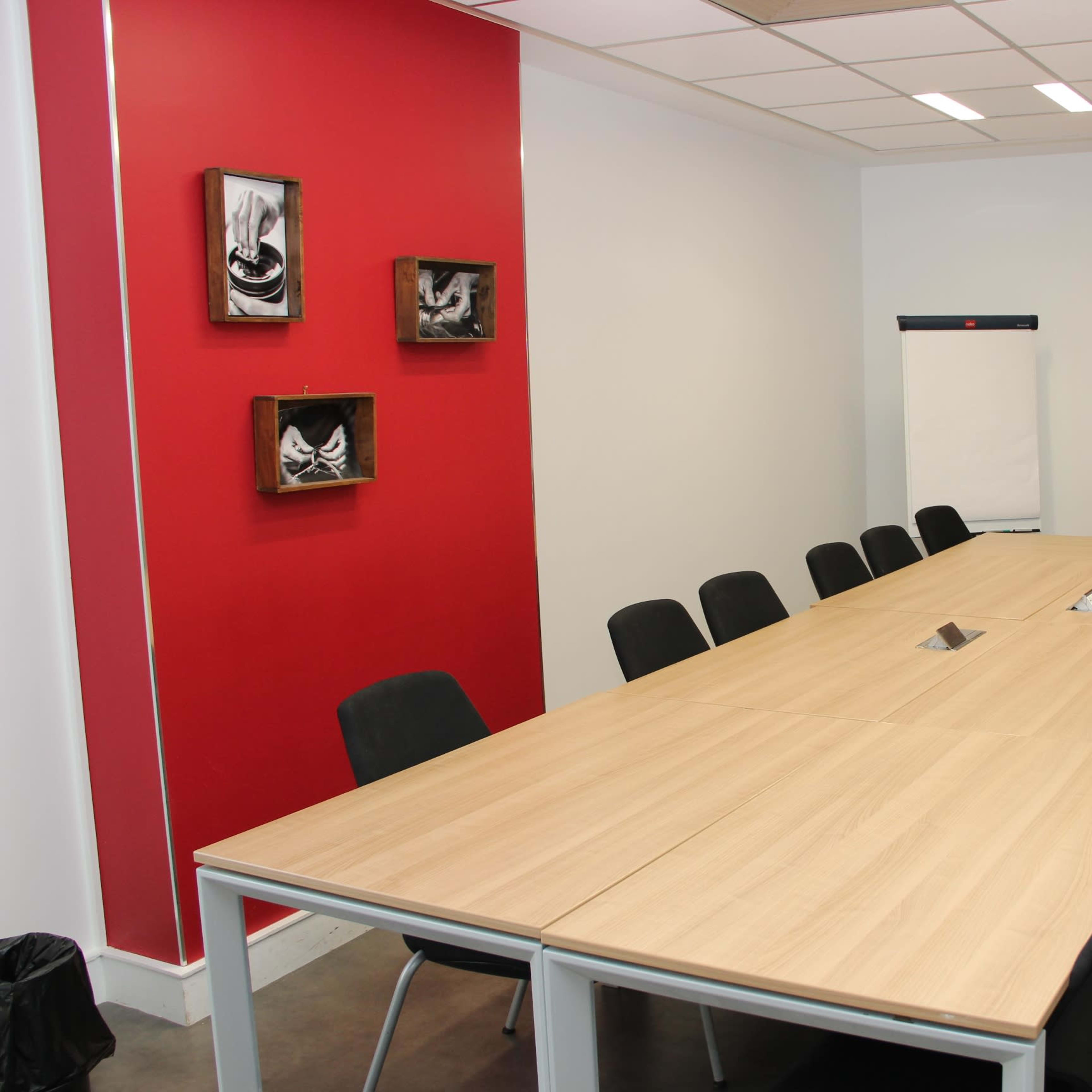 A modern conference room with a long wooden table surrounded by black chairs, featuring red accent walls and a whiteboard at one end.