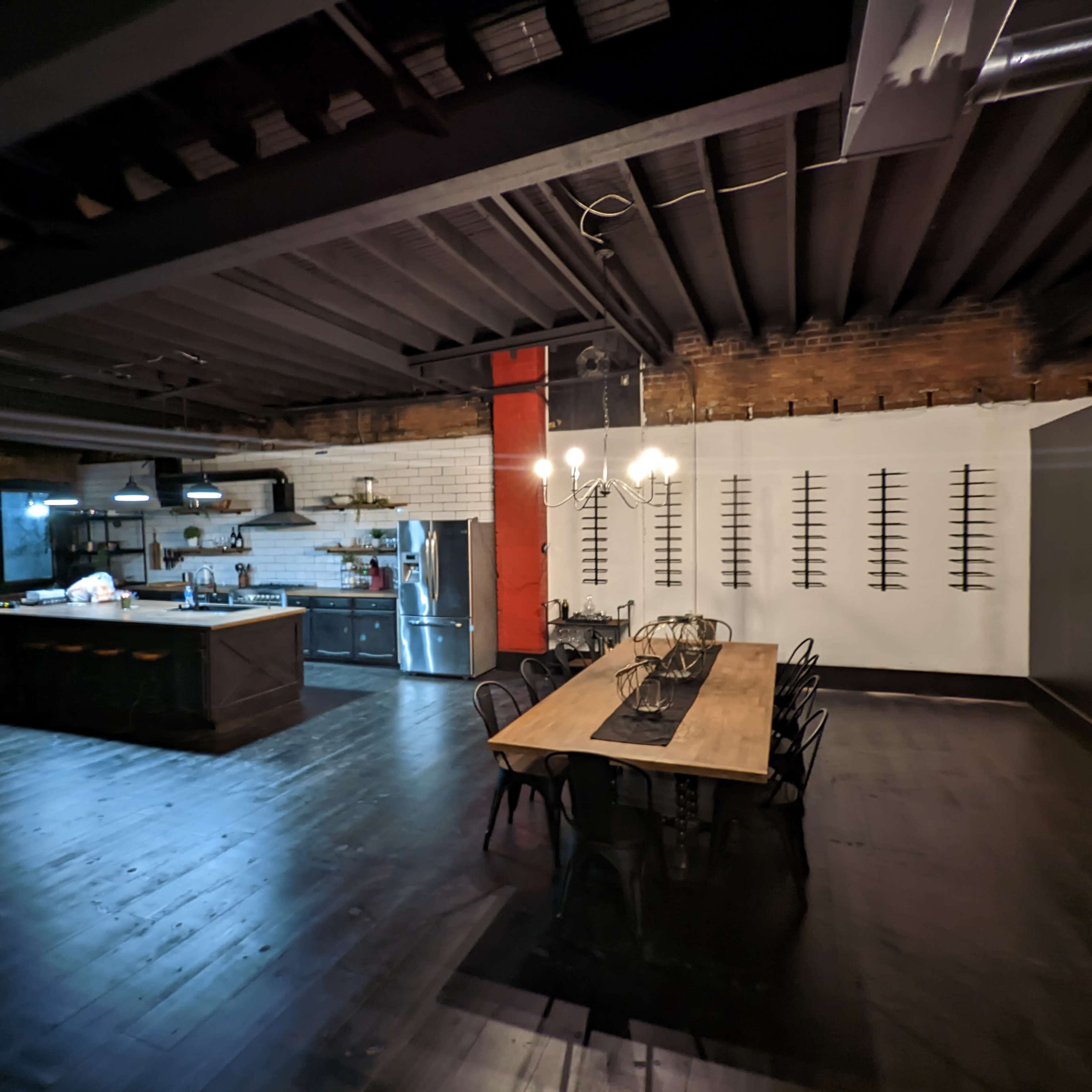 A spacious industrial-style kitchen and dining area with a long wooden table, exposed brick walls, and a black ceiling.