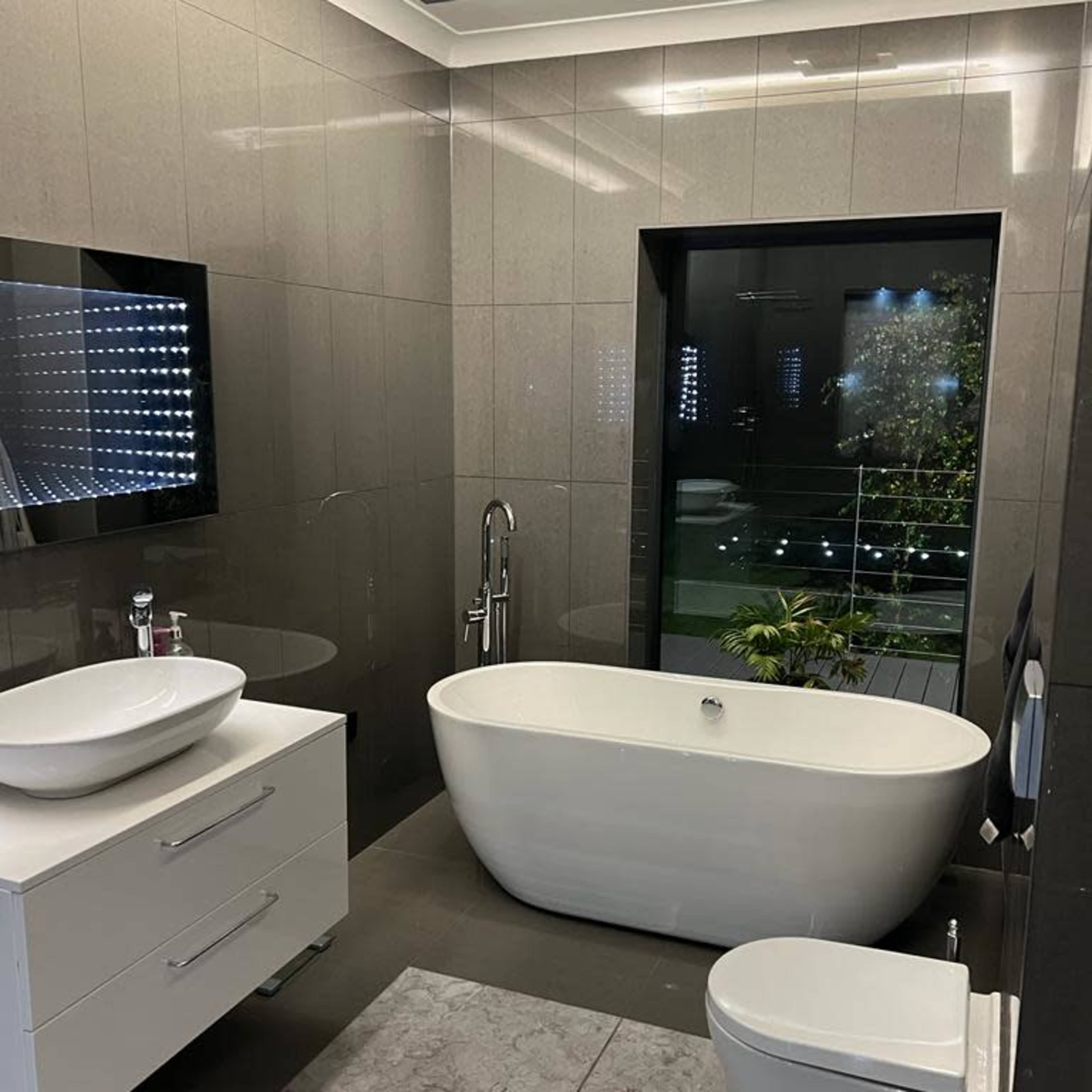 A modern bathroom featuring a freestanding bathtub, a sleek vanity with a sink, a toilet, and large windows revealing an outdoor area with greenery.