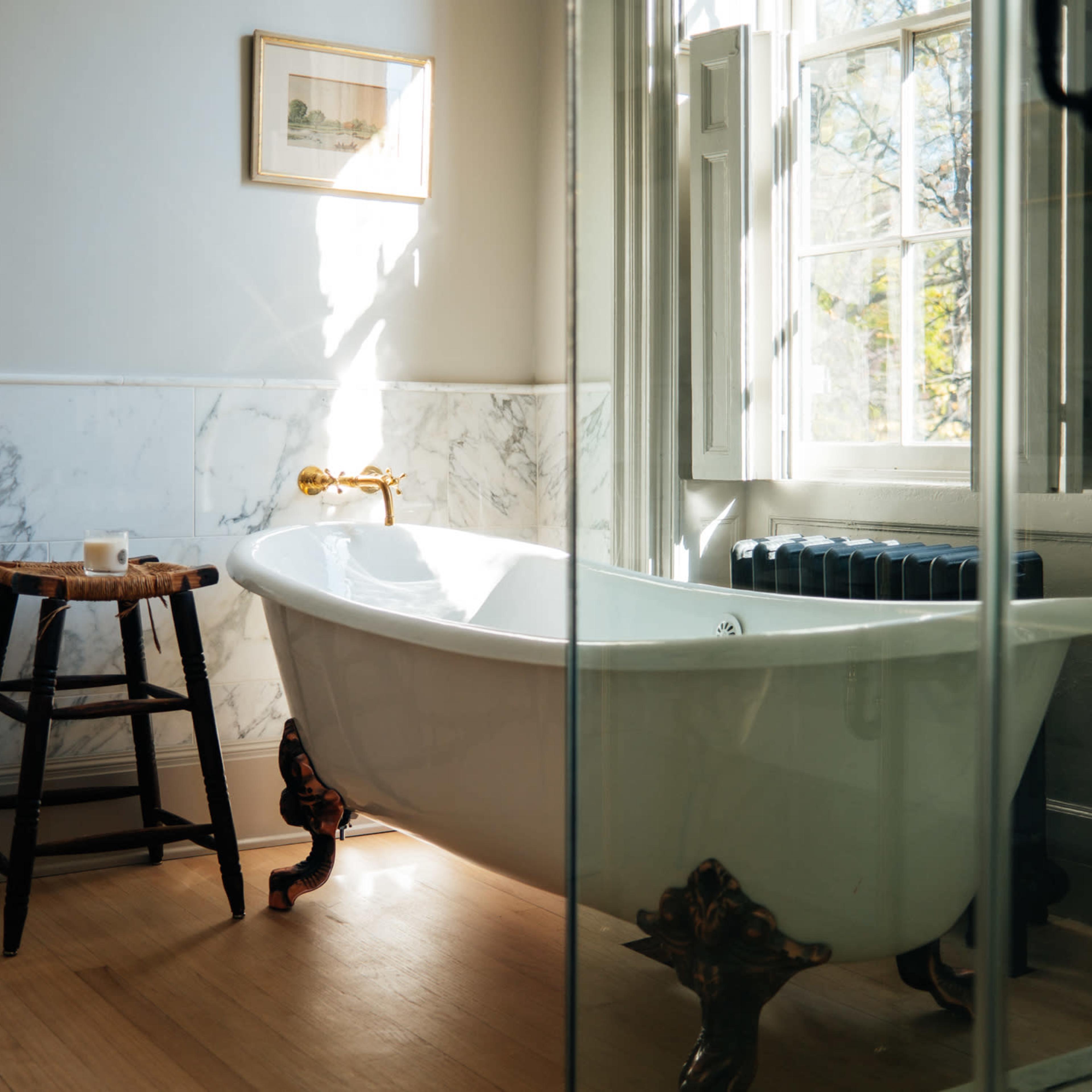 A bright bathroom featuring a clawfoot bathtub next to a wooden stool, with a window allowing natural light to enter the space.