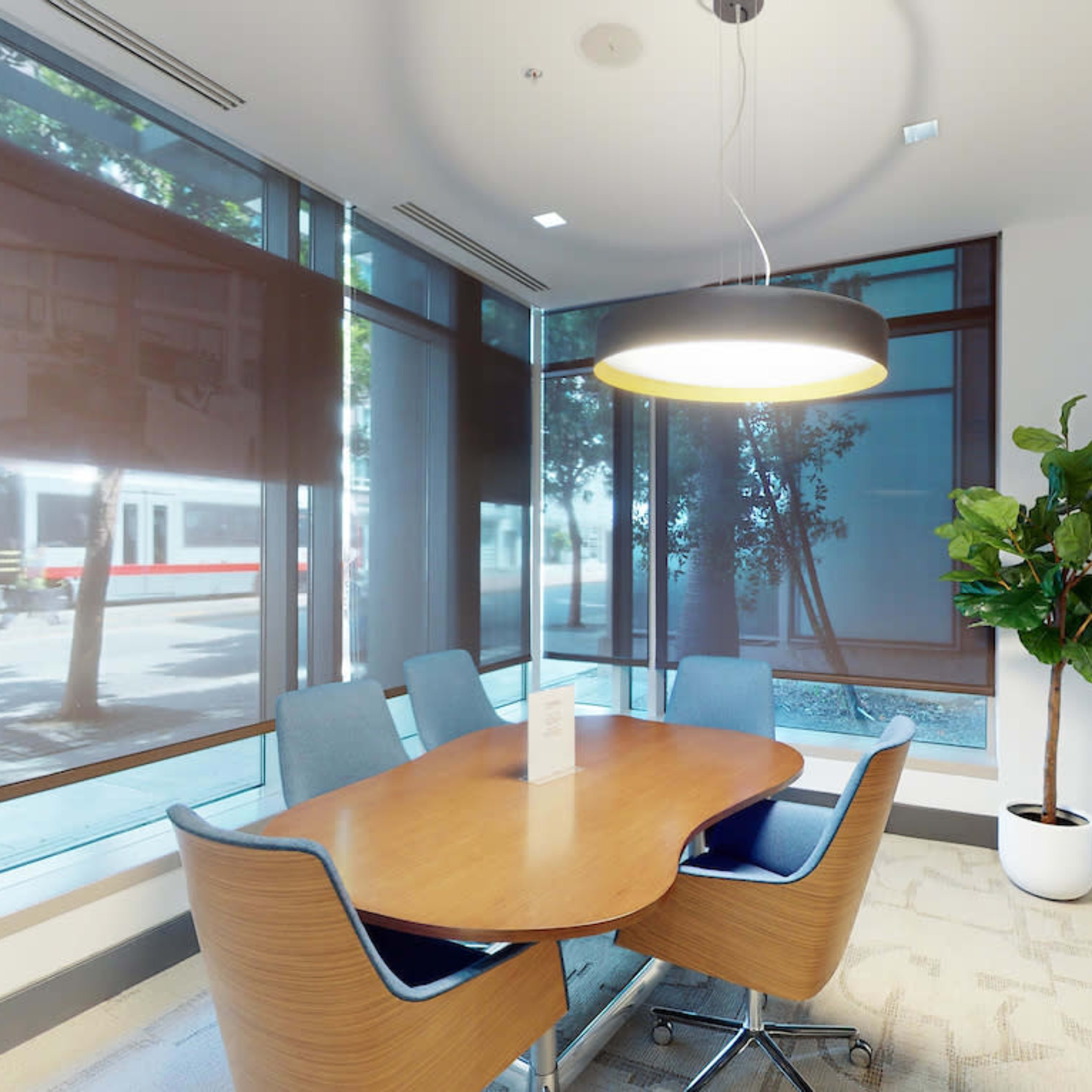 A modern conference room with a wooden table and blue chairs, featuring large windows with shades and a decorative plant in the corner.