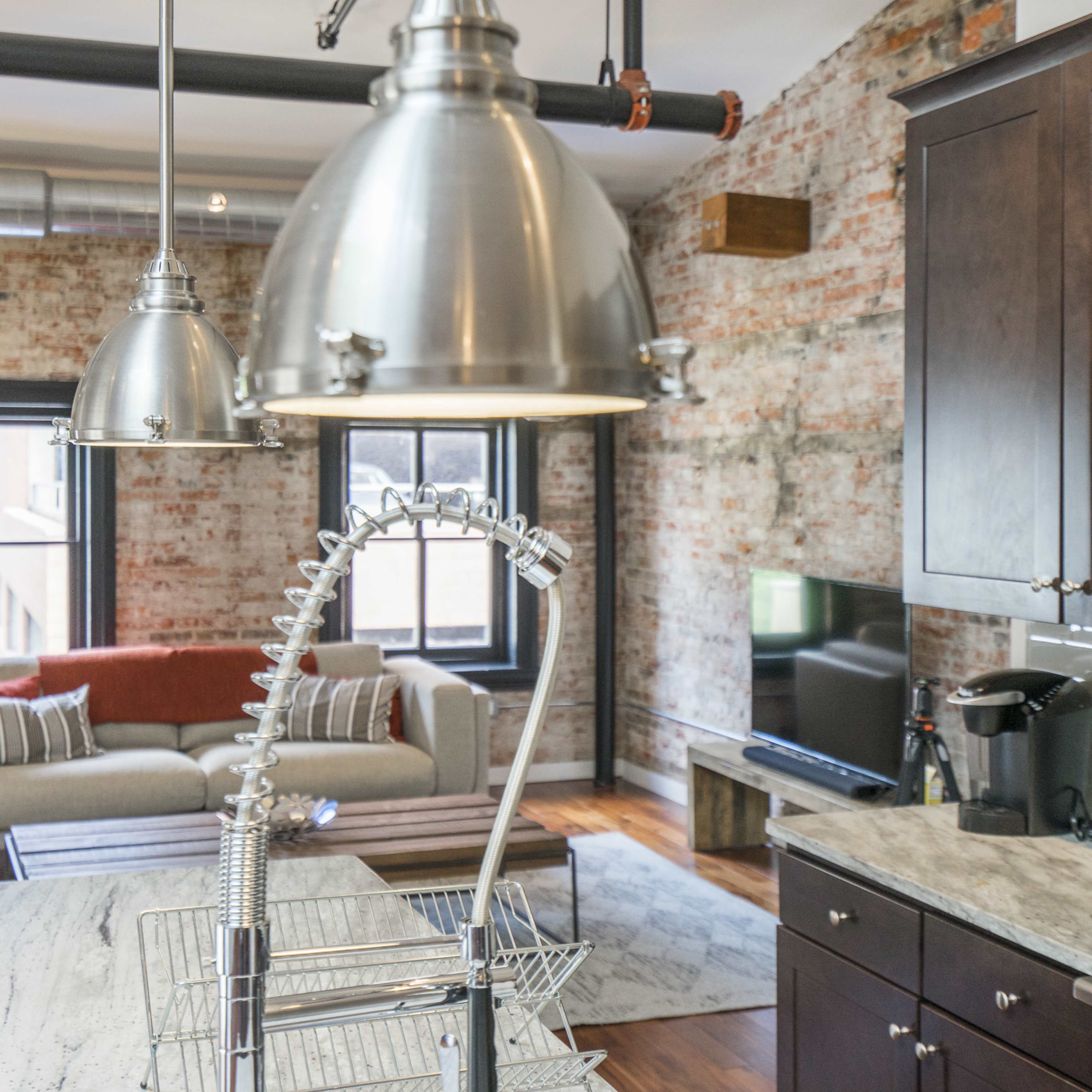 A modern kitchen with a large island, industrial-style pendant lights, and brick walls, adjacent to a cozy living area with a sofa and television.