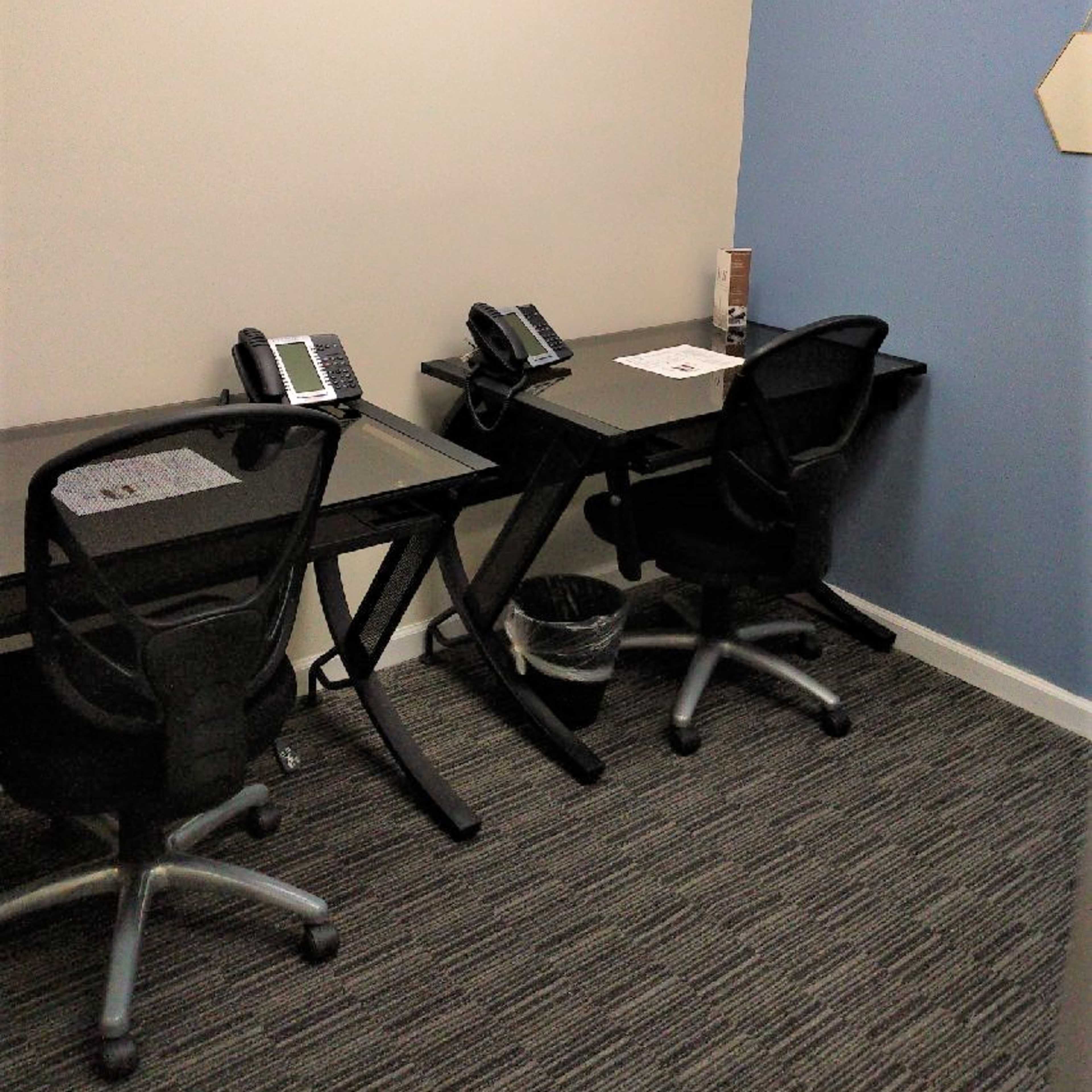 Two black desks with phones and chairs set against a light-colored wall in an office corner.