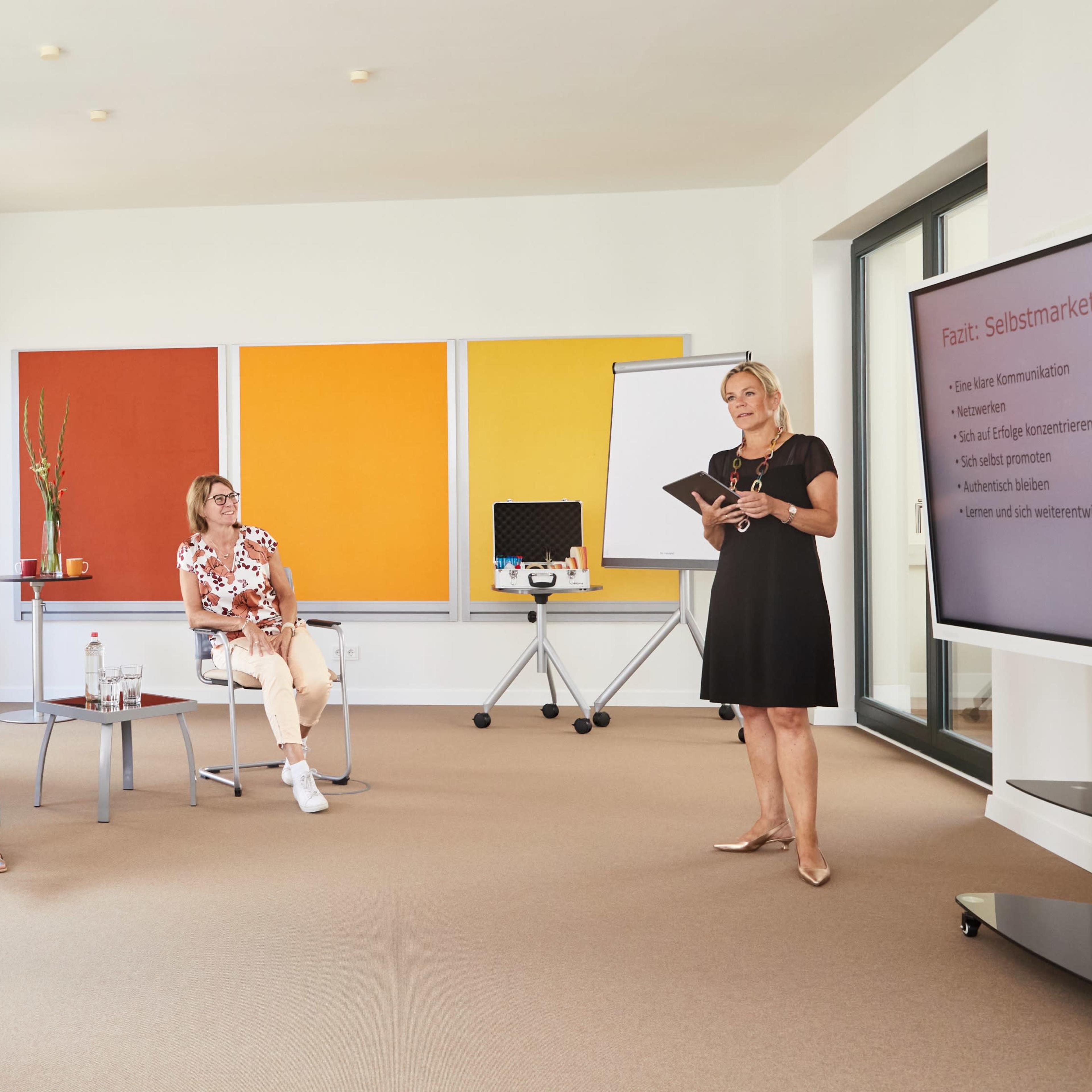 A group of four women sit in a conference room while another woman presents information on a screen.