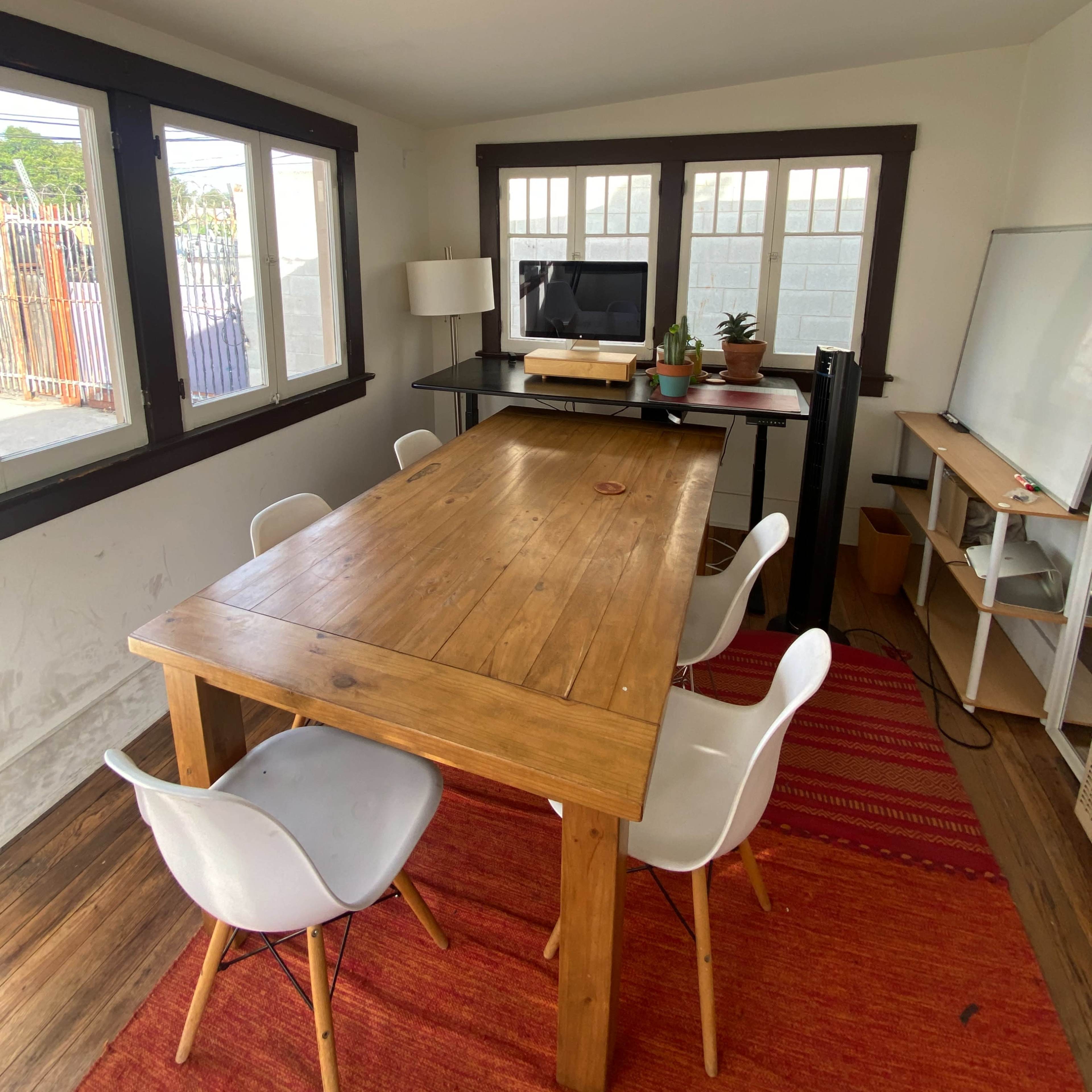 A wooden table surrounded by white chairs is positioned in a well-lit room with large windows, a television, and a whiteboard.