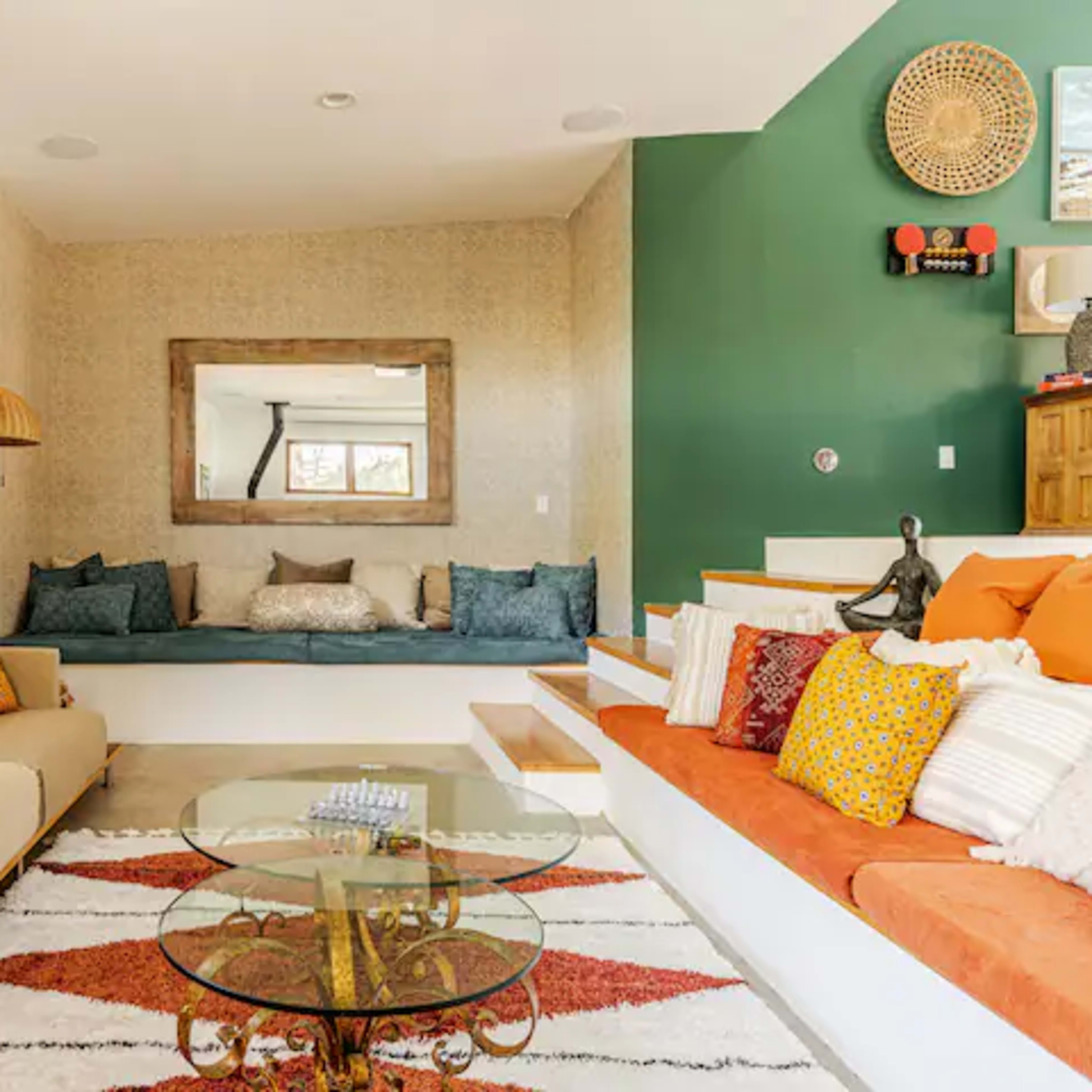 A modern living room featuring a beige sofa, a glass coffee table, a colorful area rug, and a seating area with cushions against the wall, complemented by decorative wall art and a mirror.