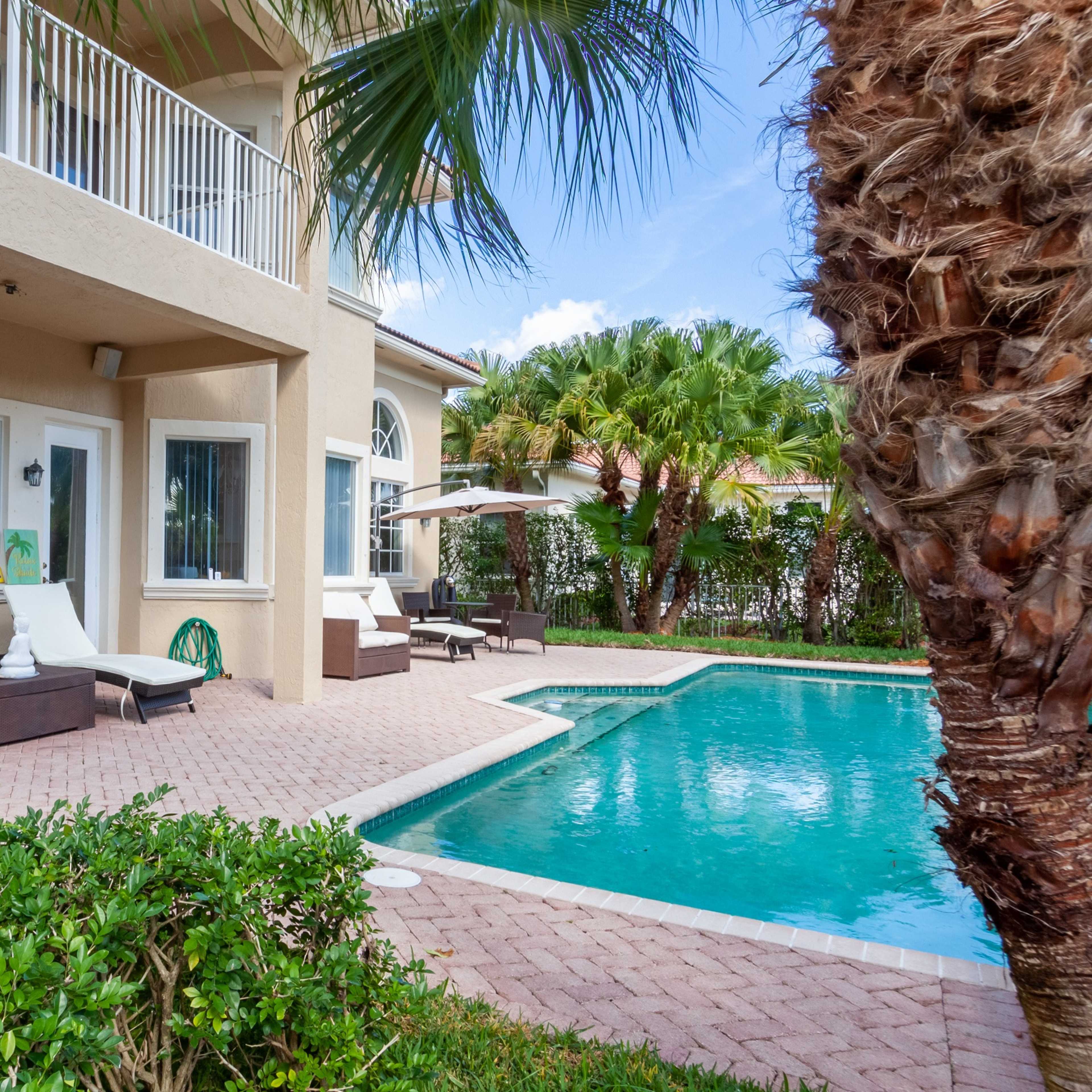 A two-story house with a swimming pool and lounge chairs sits in a landscaped yard surrounded by palm trees.
