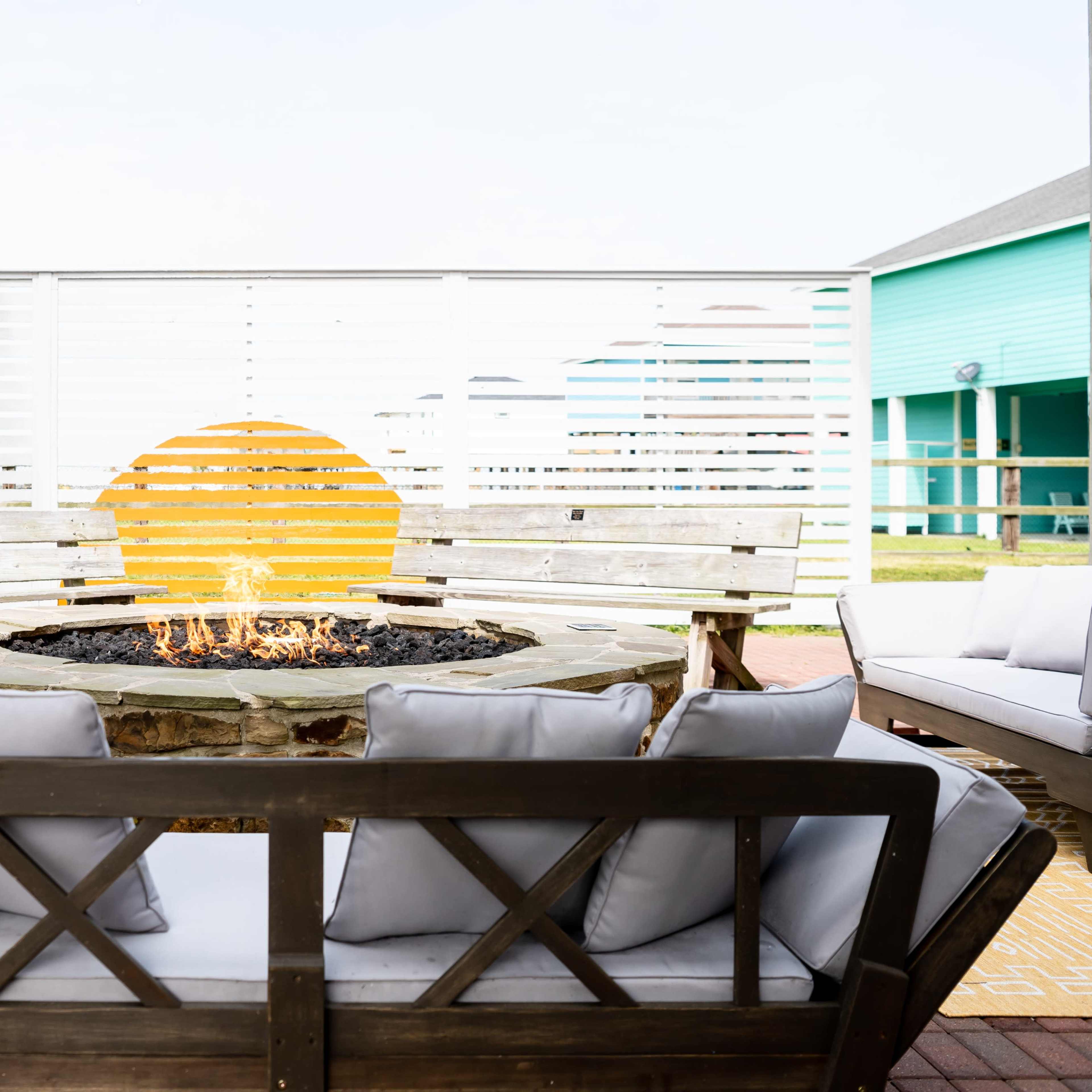 A fire pit surrounded by wooden benches and cushions is set against a backdrop of a white slatted wall with a yellow circular design, indicating sunset.