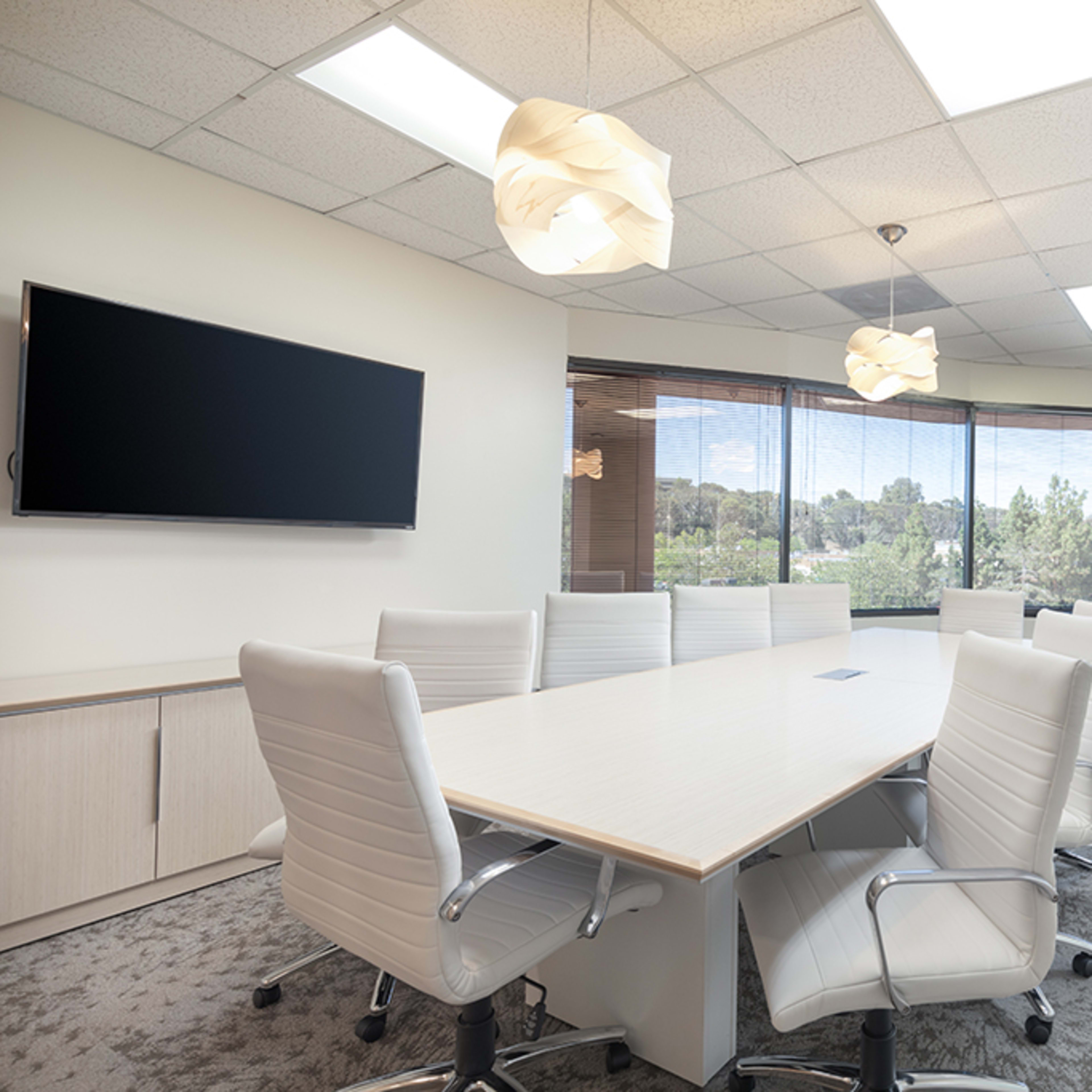 A modern conference room features a large table surrounded by white chairs, with a wall-mounted screen and windows overlooking greenery.