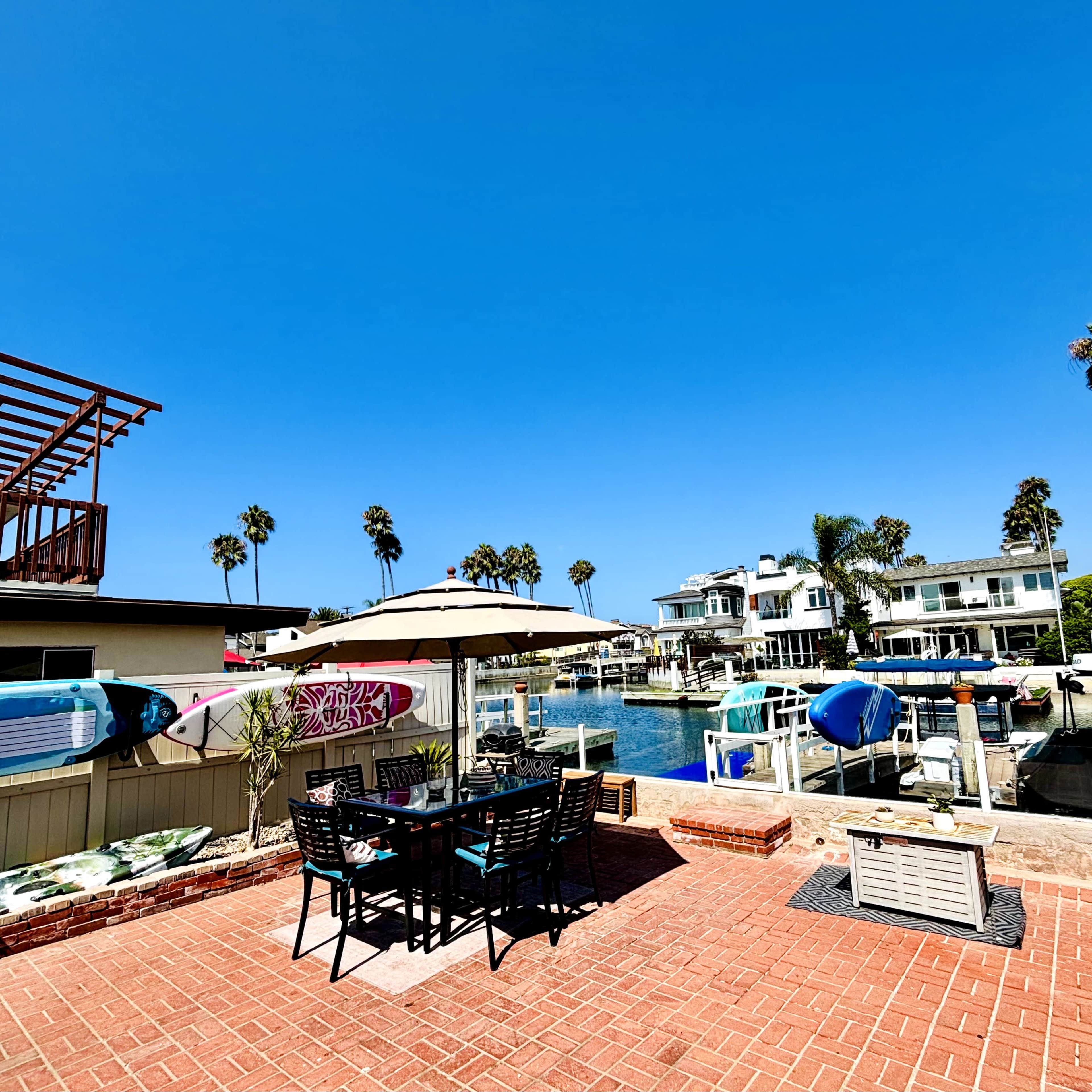 A patio with a dining set overlooks a calm canal lined with palm trees and waterfront homes.
