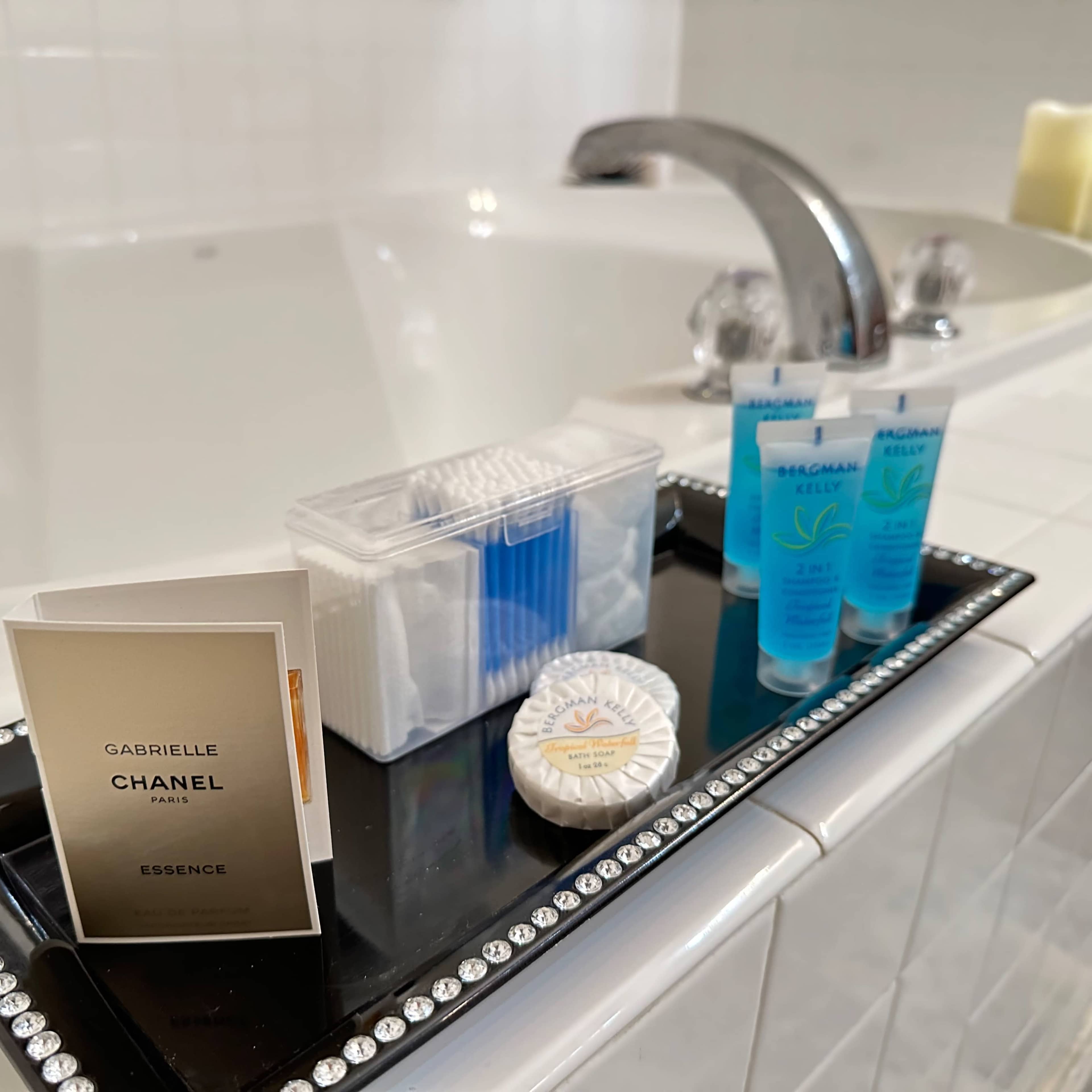 A tray holds a Chanel perfume sample, cotton swabs, a small package of cotton pads, and two tubes of moisturizer near a bathtub.