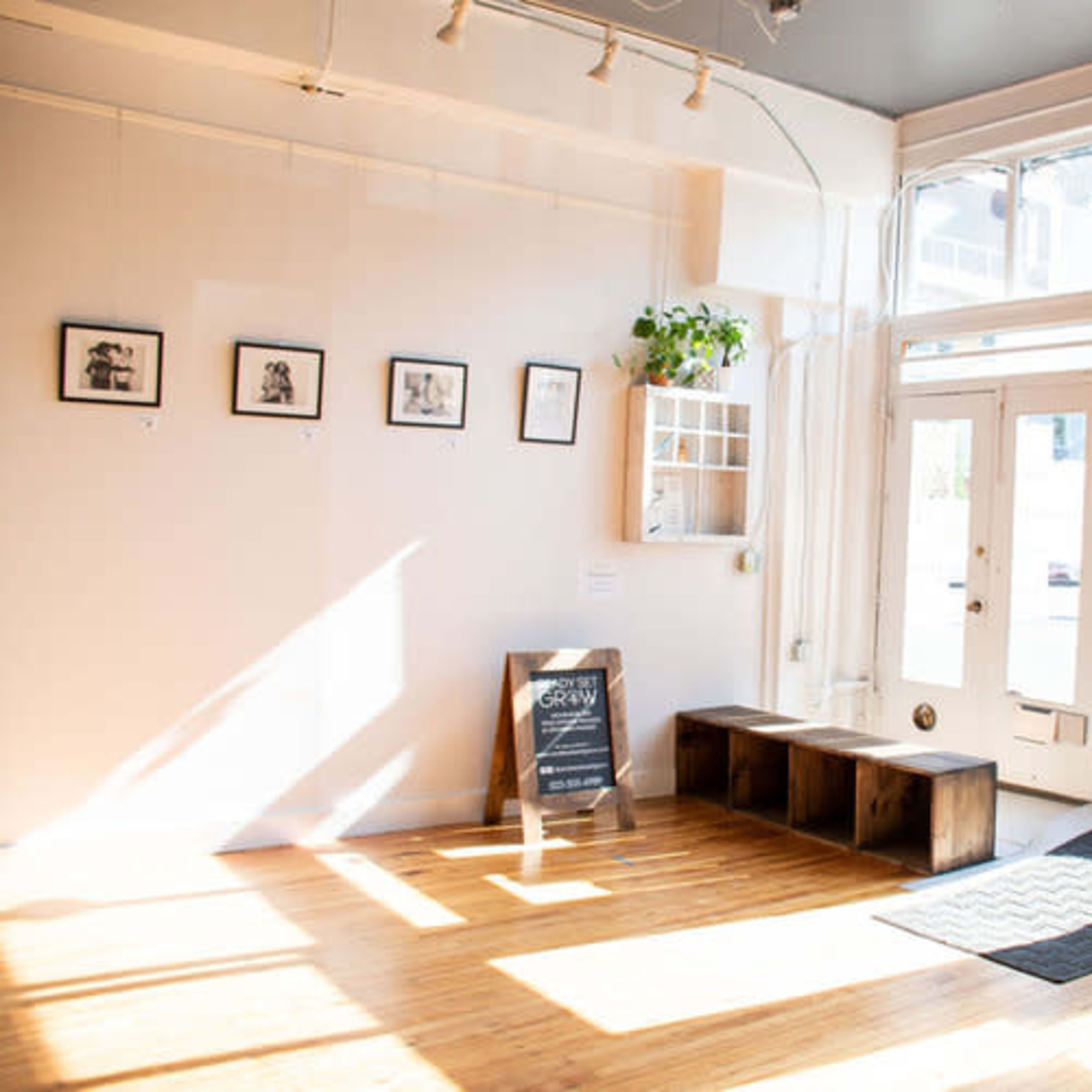 A bright, sparsely furnished room features wooden benches, framed pictures on the wall, and large windows allowing natural light to enter.