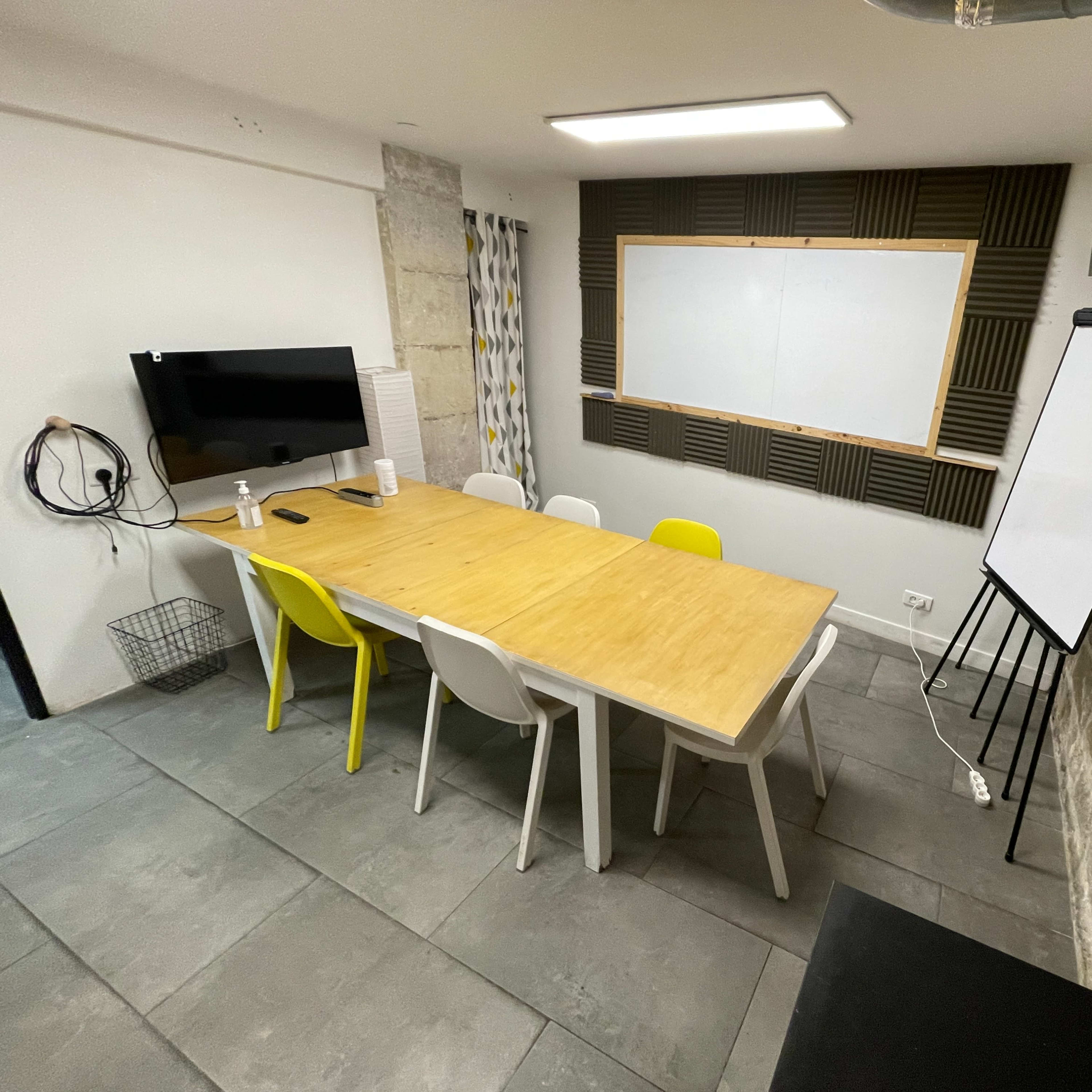 A meeting room features a long wooden table surrounded by colorful chairs, a whiteboard, a television on the wall, and exposed stone elements.