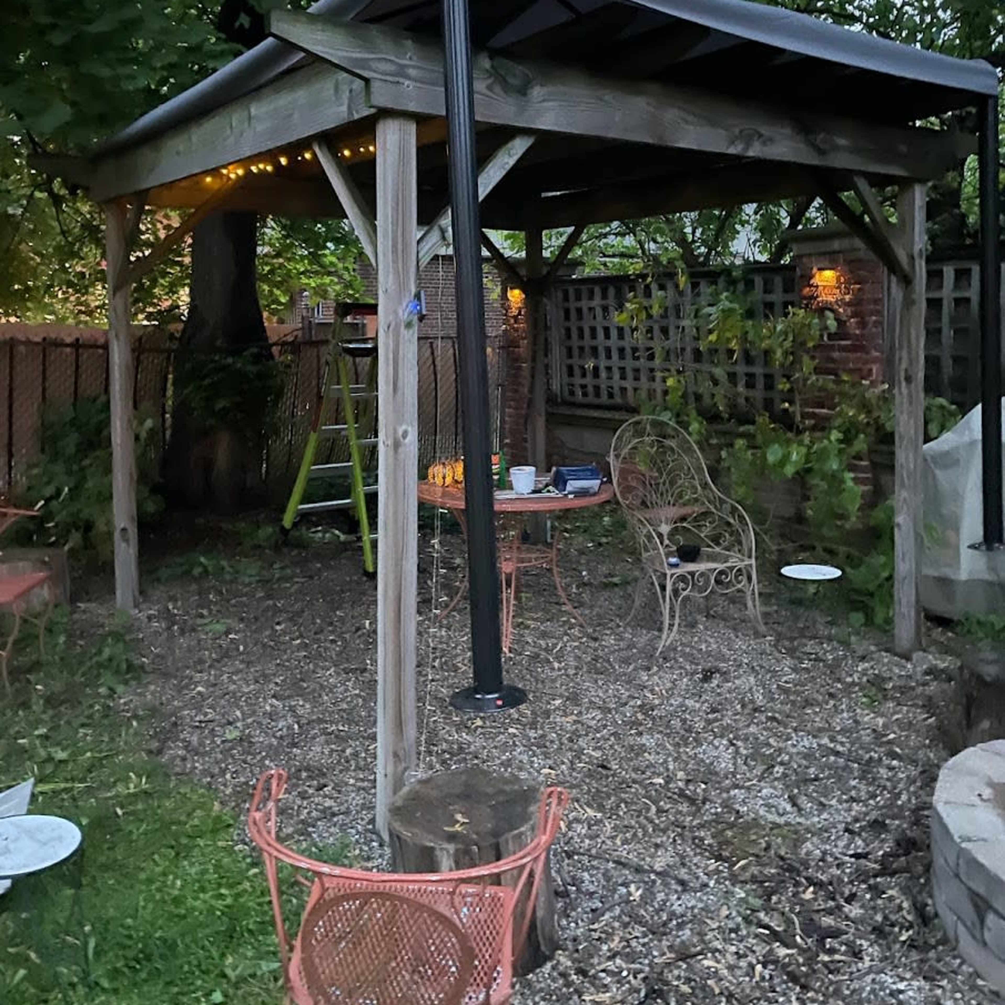 A wooden gazebo in a backyard, surrounded by trees and featuring outdoor furniture and a ladder nearby.
