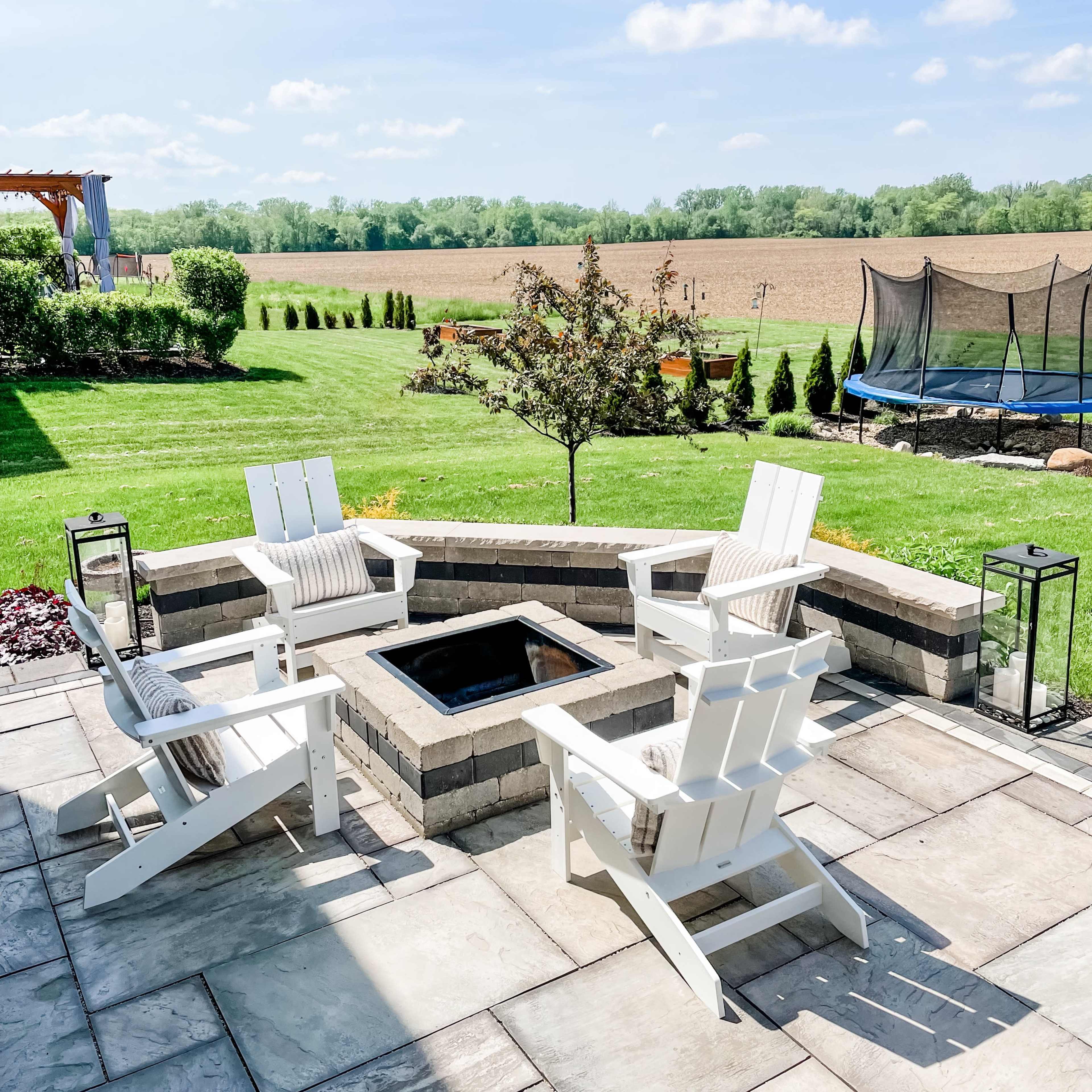 A patio area with four white Adirondack chairs arranged around a square stone fire pit, surrounded by a green lawn and a trampoline in the background.