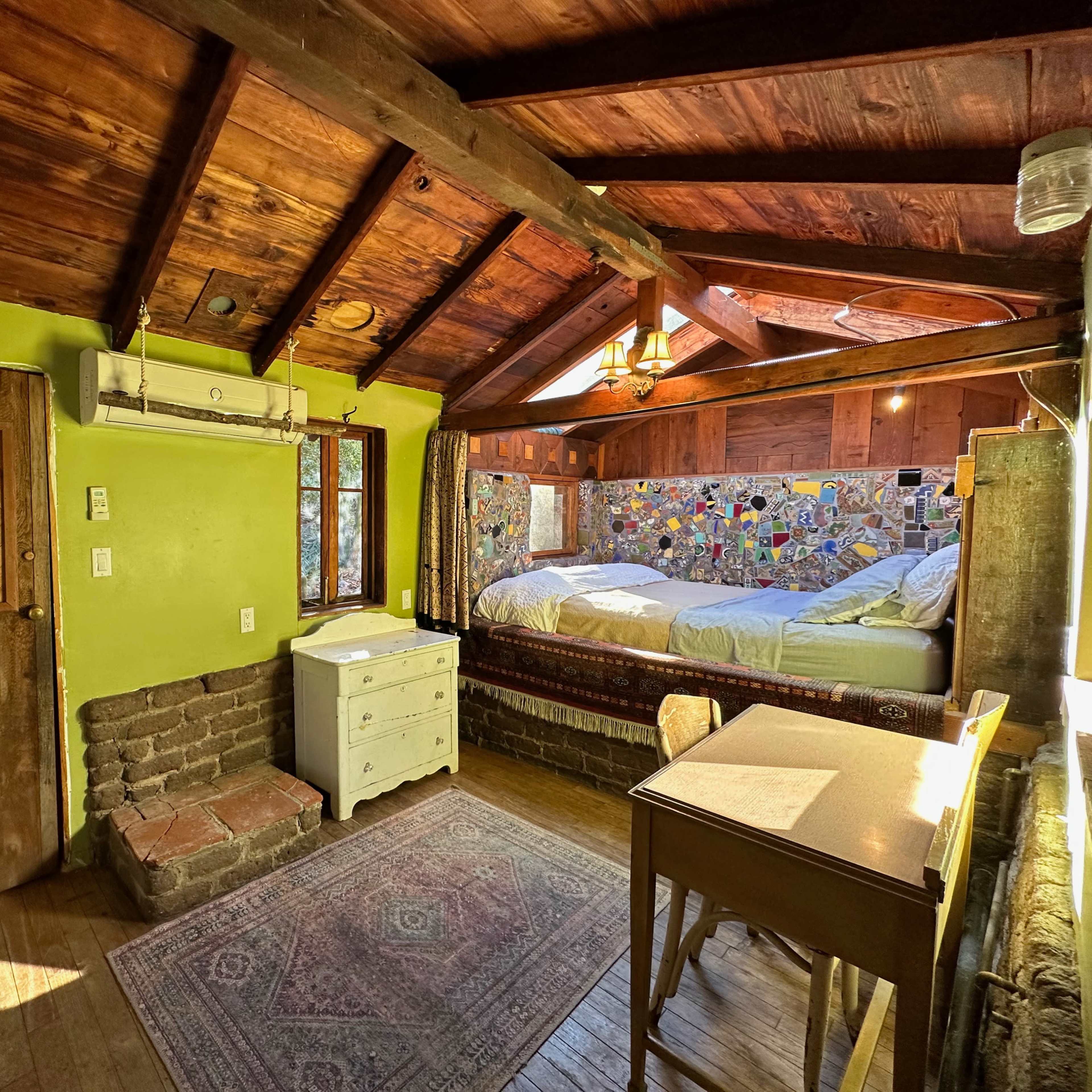 A cozy room with a wooden ceiling, a double bed with a colorful mural behind it, a small desk, and a door leading outside.