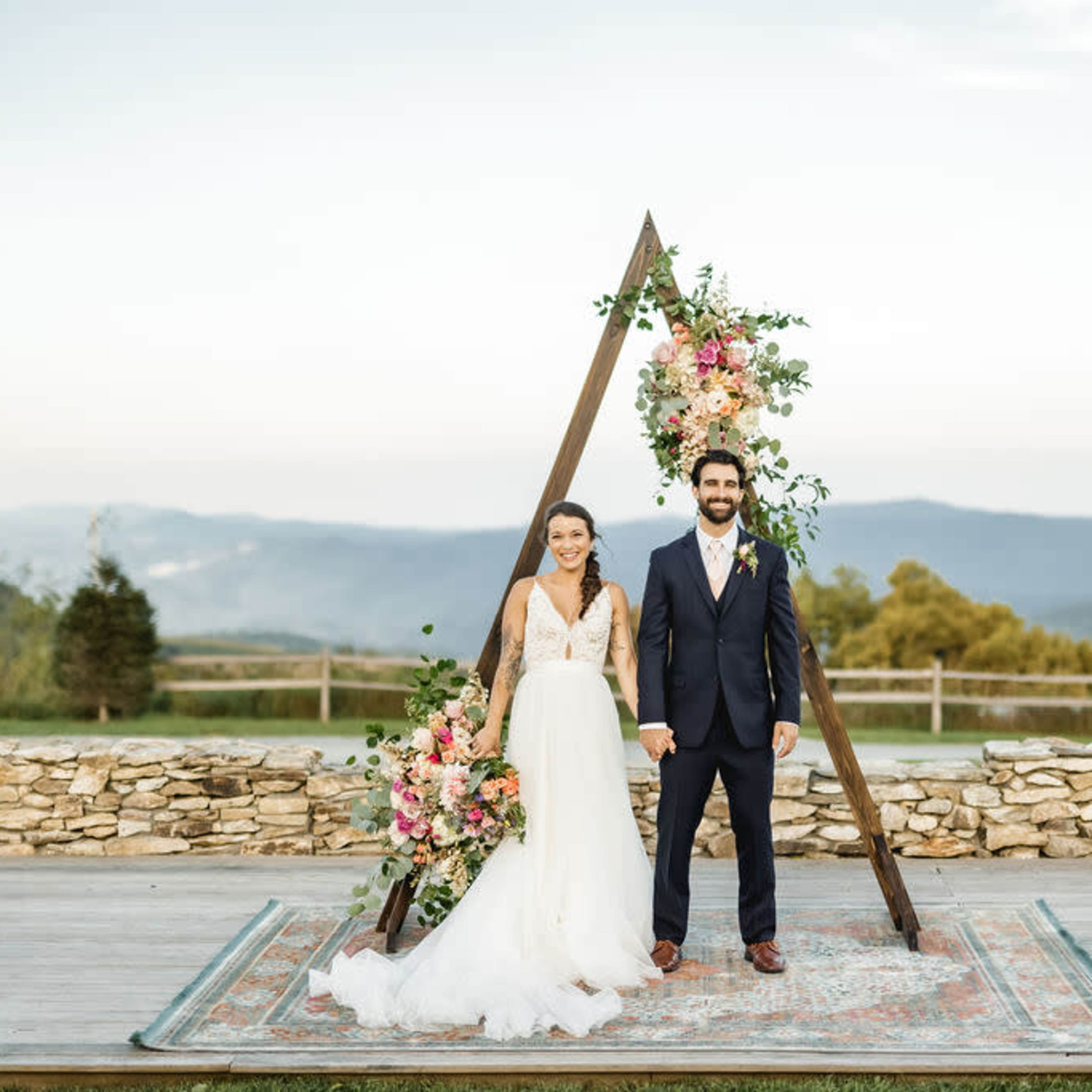 A bride and groom stand in front of a floral-decorated wooden arch with mountains in the background.