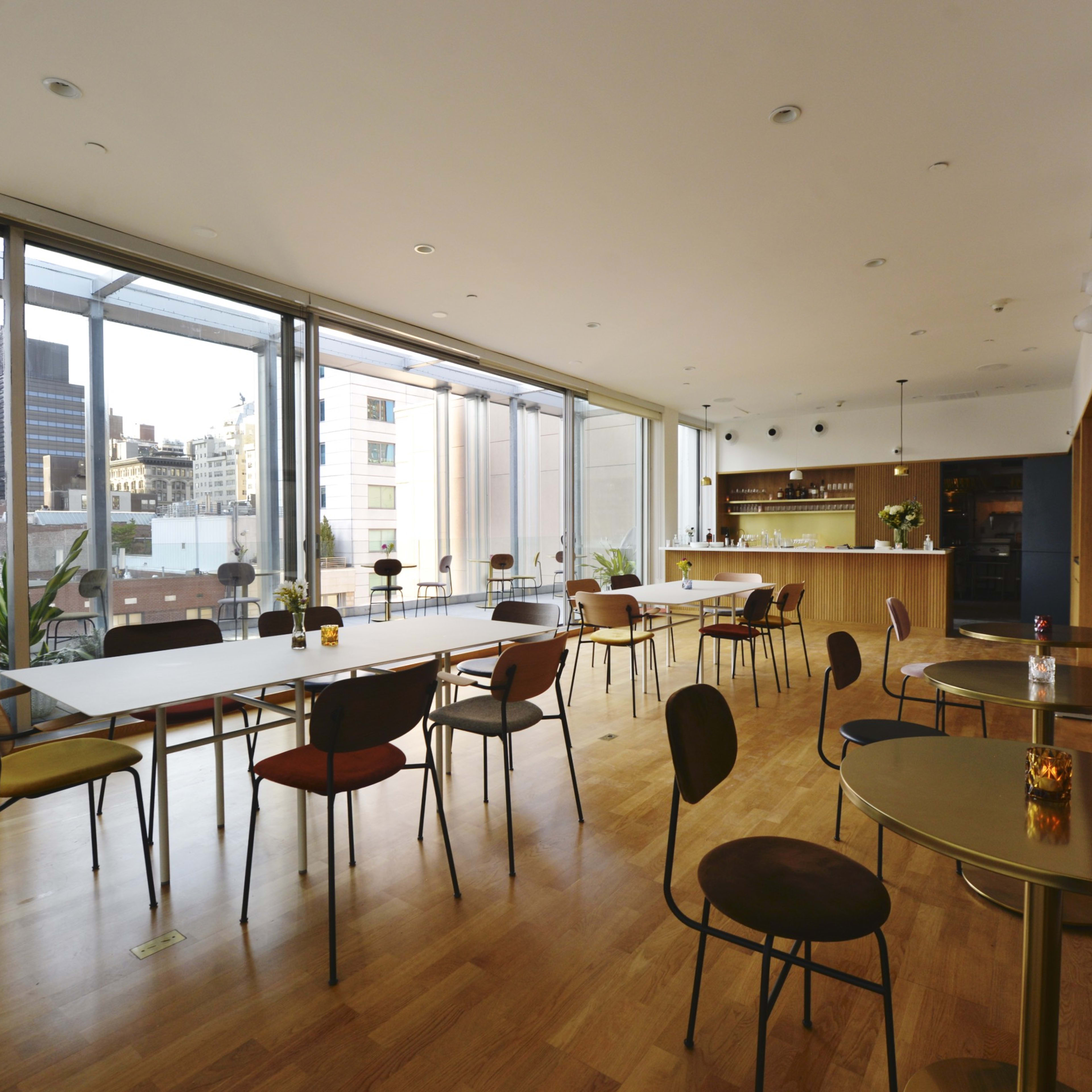 A modern, spacious interior of a cafe or restaurant, featuring a long table, various chairs, and large windows that provide a view of the city skyline.