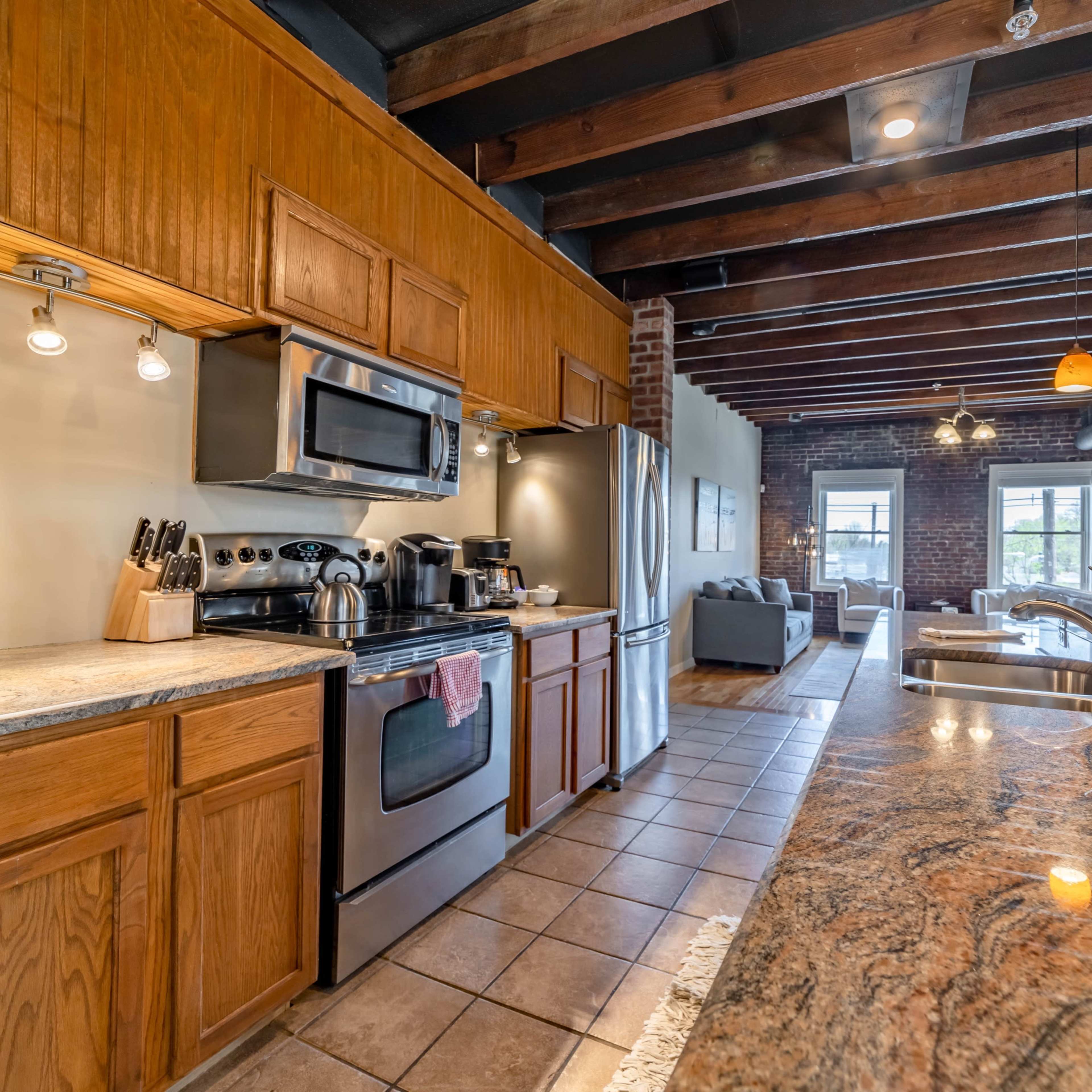 A modern kitchen with wooden cabinetry, stainless steel appliances, and an open layout leading to a living area with exposed brick walls.
