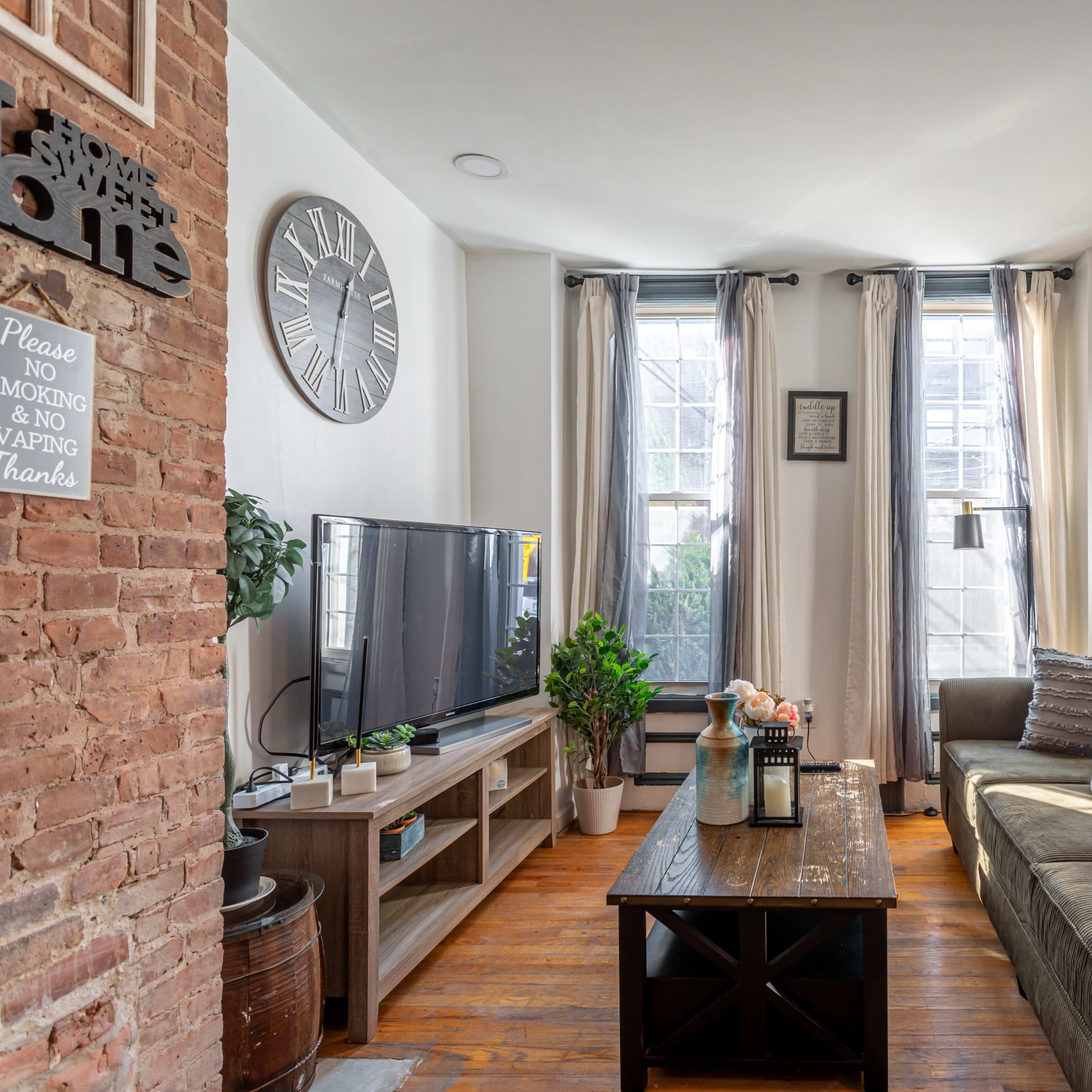 A cozy living room with a large sofa, a television, and plants, featuring natural light coming through two windows.