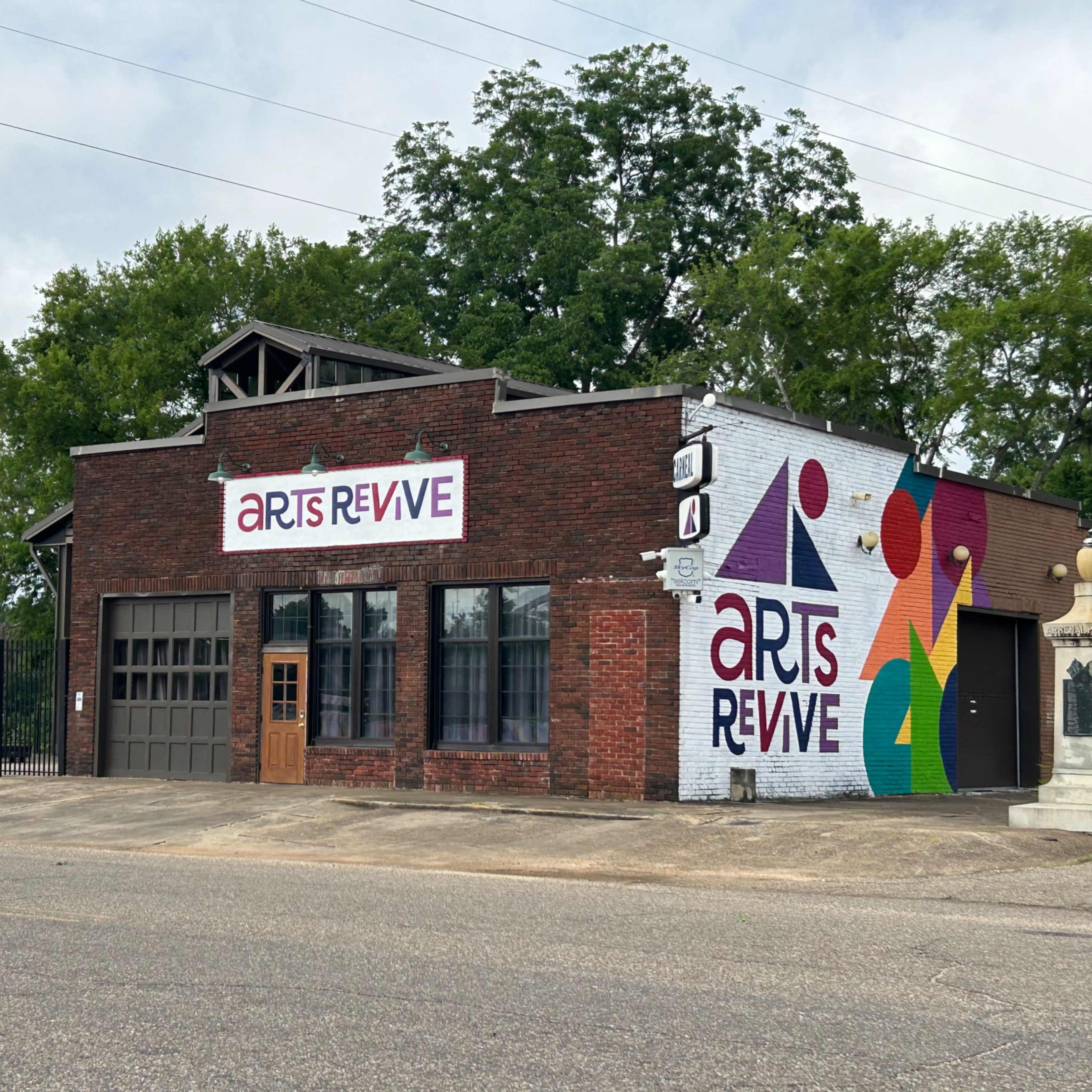 A brick building with large "Arts Revive" signage on the front, adjacent to a fenced area and surrounded by trees.