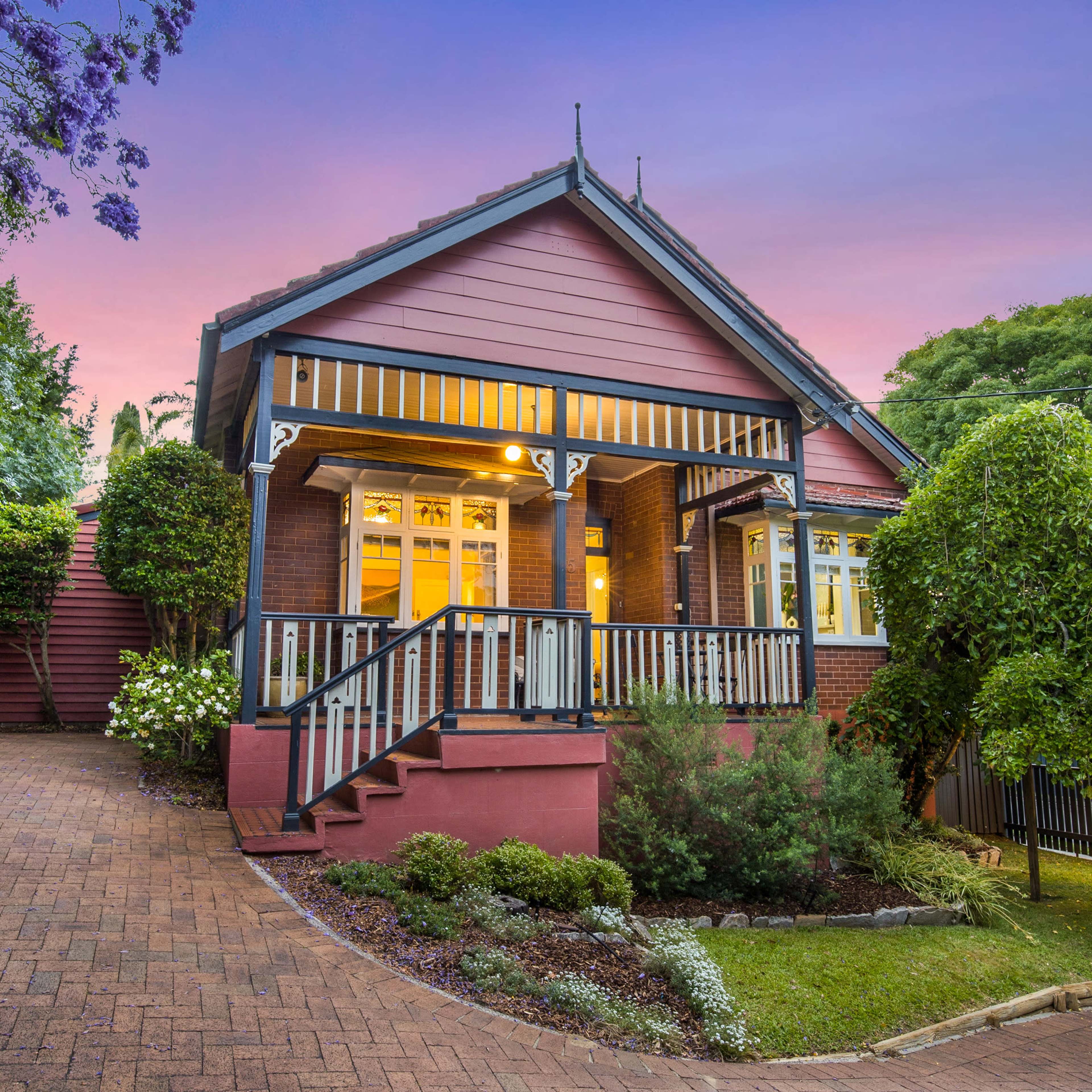 A two-story house with a front porch is surrounded by greenery and shrubbery, with a brick driveway leading up to it.