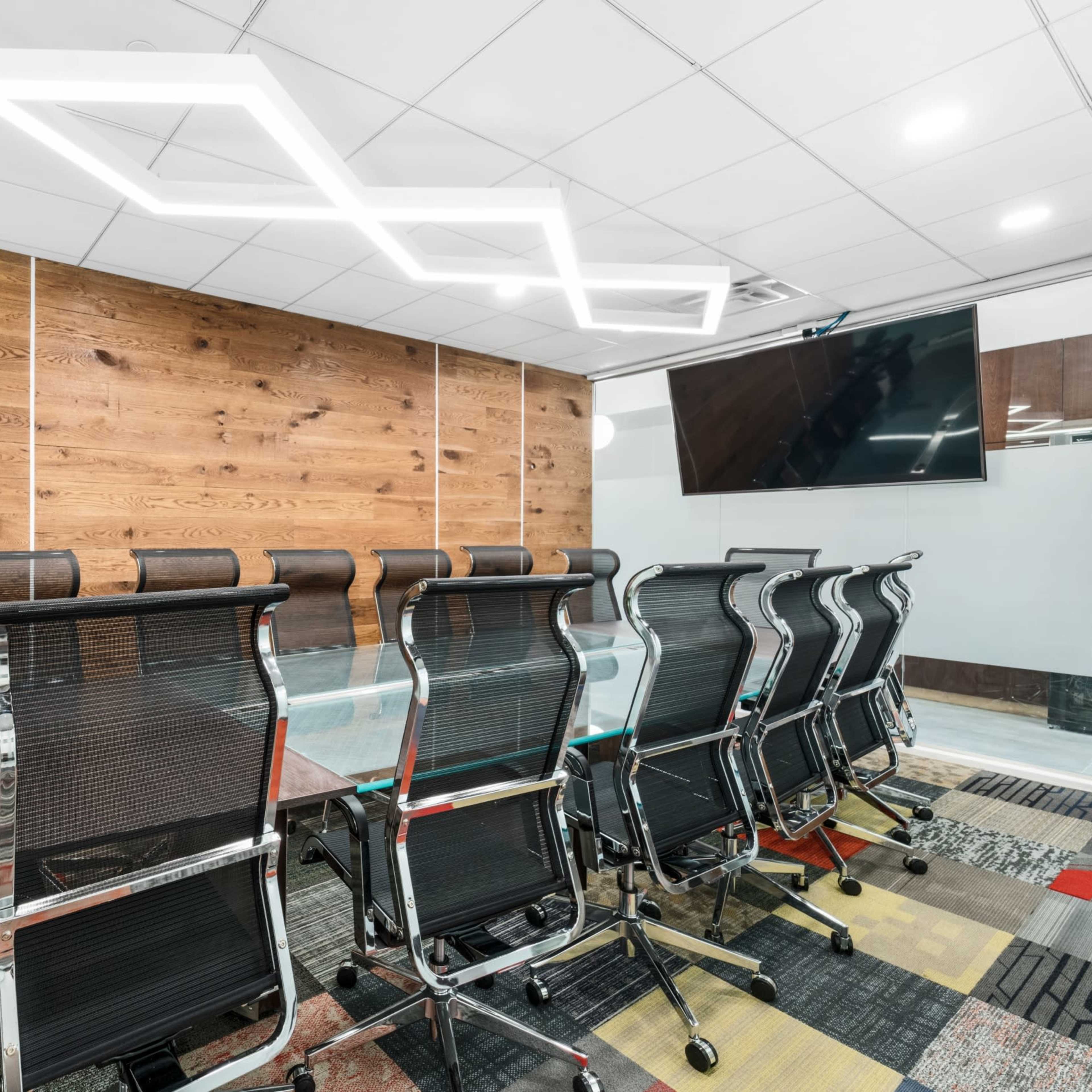 A modern conference room features a long table surrounded by black ergonomic chairs, with a wooden accent wall and contemporary lighting above.