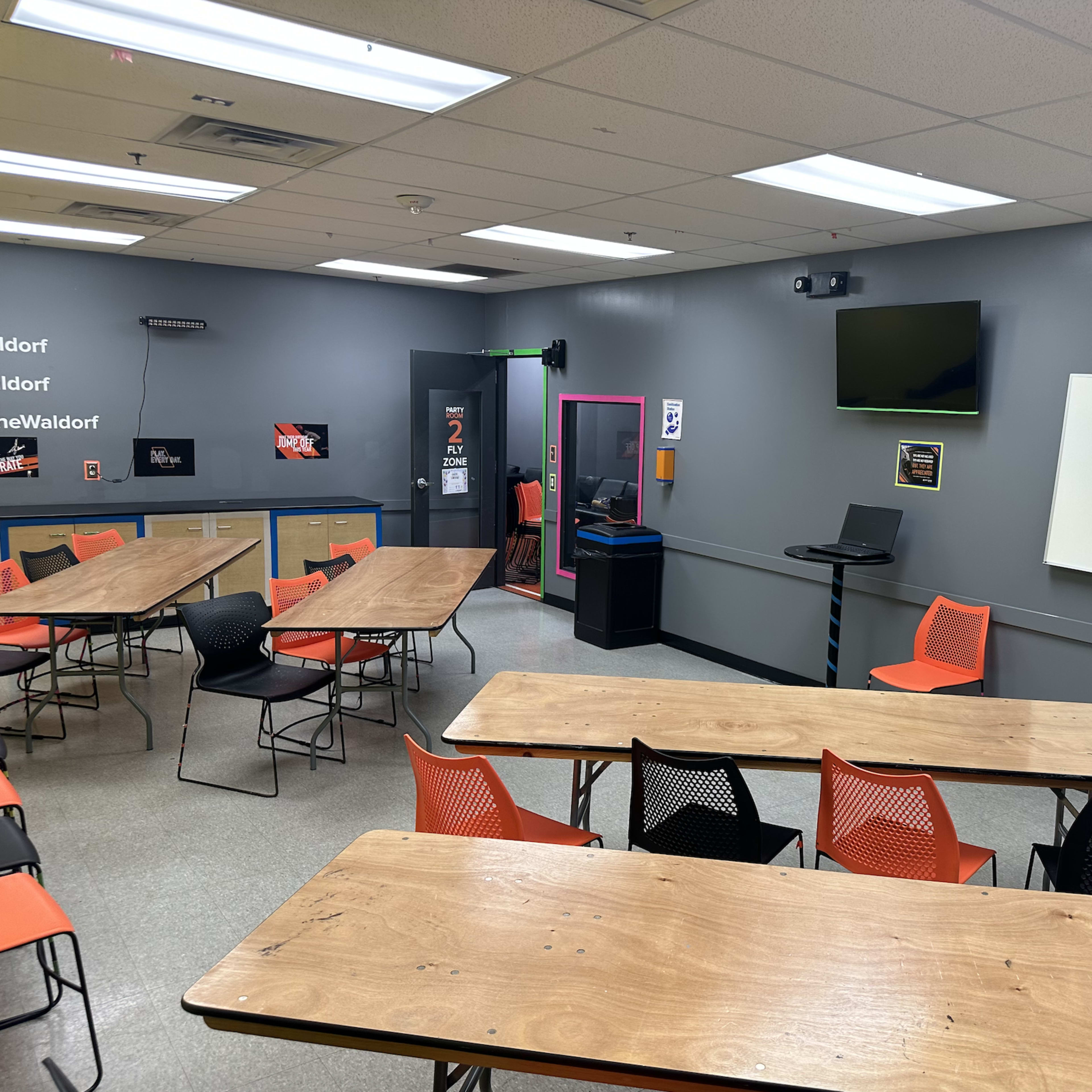 A classroom is equipped with tables, chairs, a television, a whiteboard, and a computer station against a gray wall.