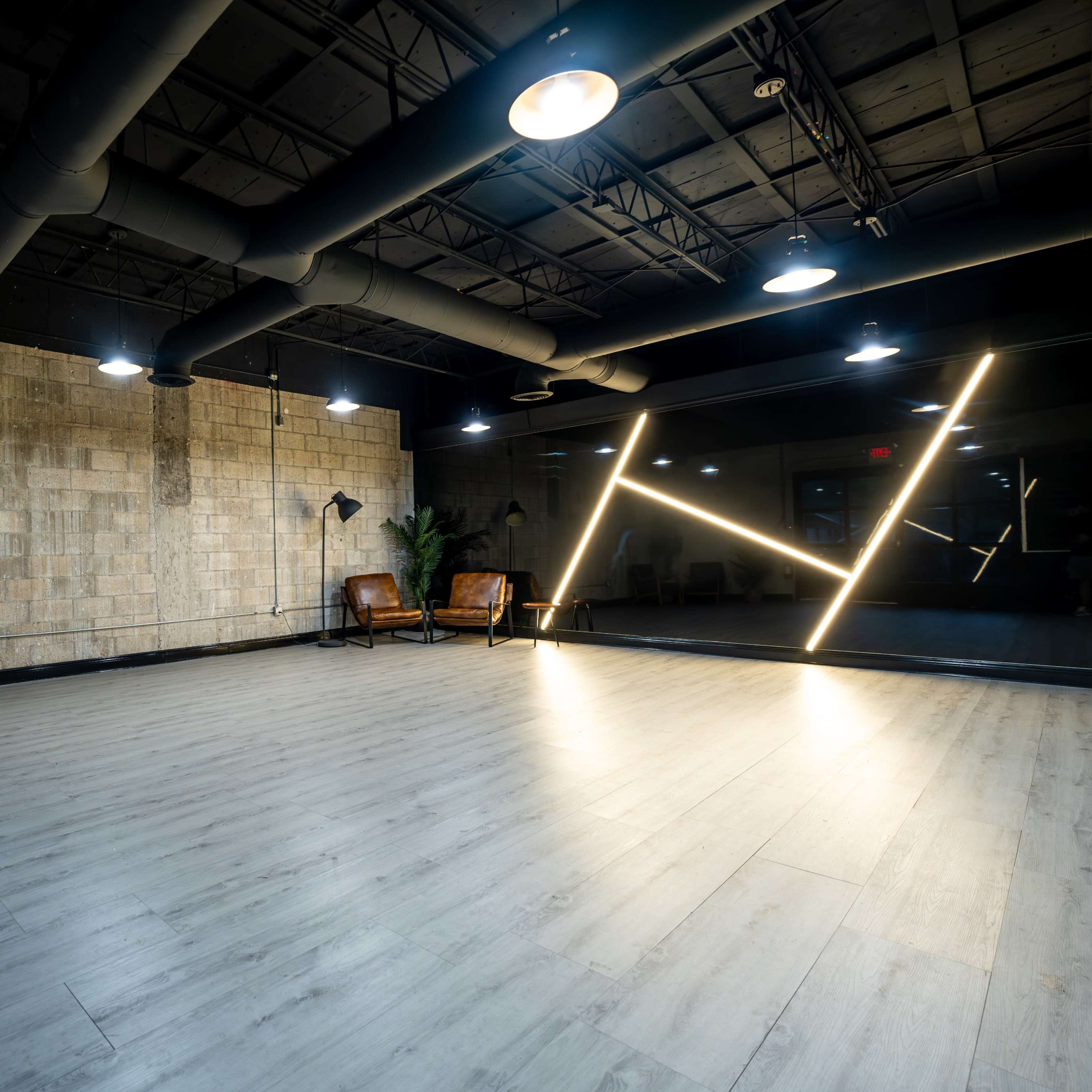 A spacious room with a light wooden floor, exposed brick walls, and minimalist furniture, illuminated by modern lights and featuring a decorative neon fixture.