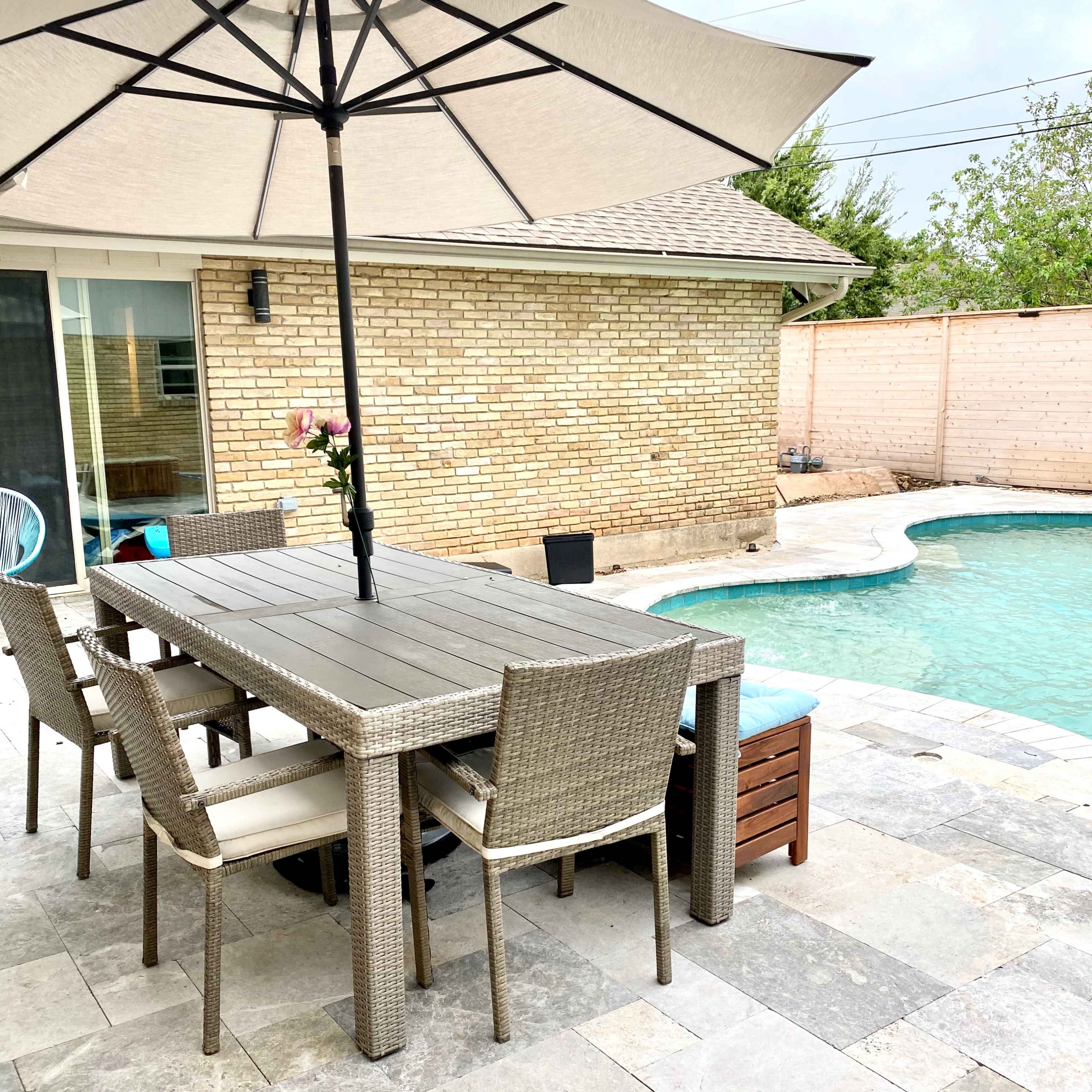 A dining table with chairs is set next to a pool, shaded by a large umbrella, in a backyard with stone paving.