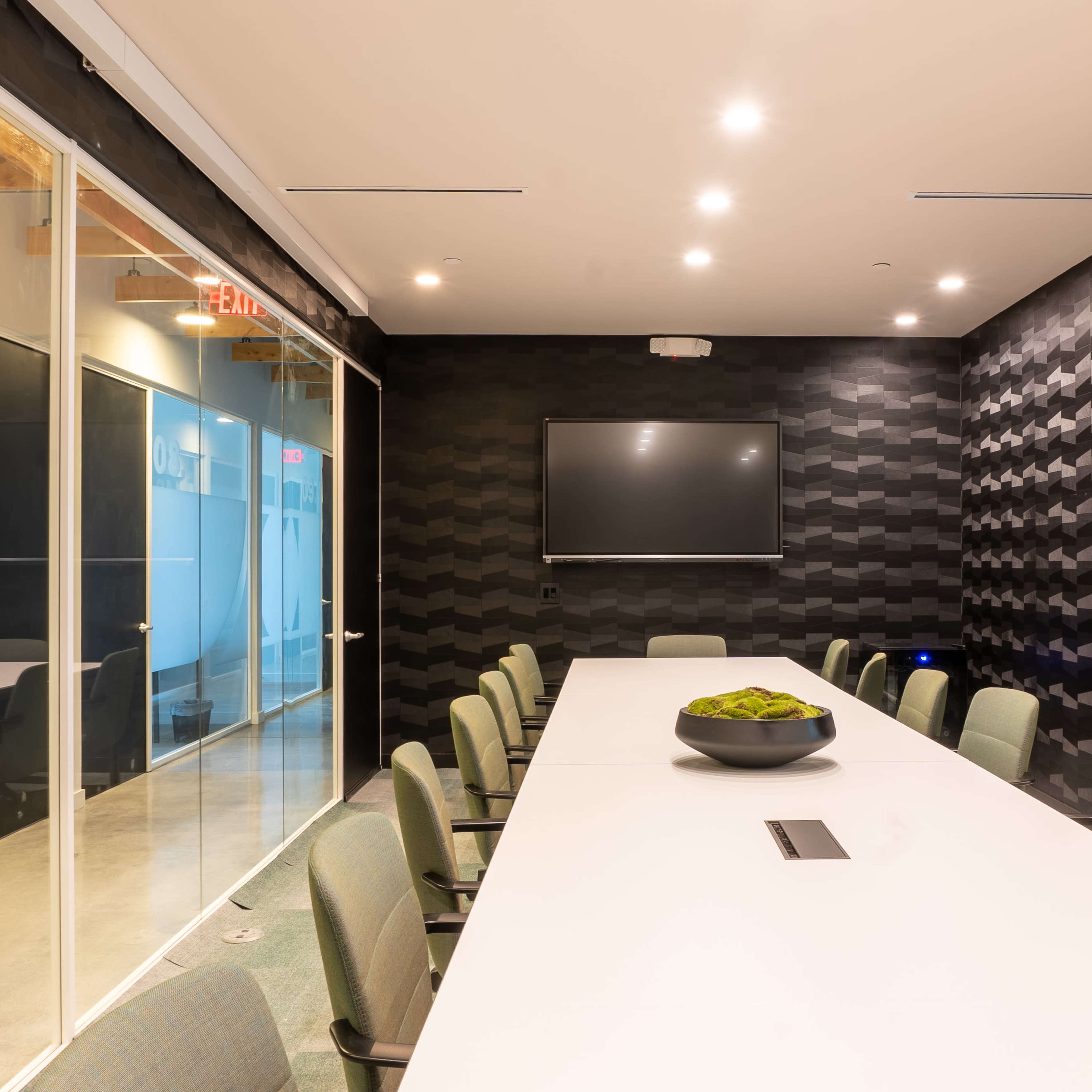A modern conference room features a long white table surrounded by green chairs, with a large screen mounted on a textured black wall.