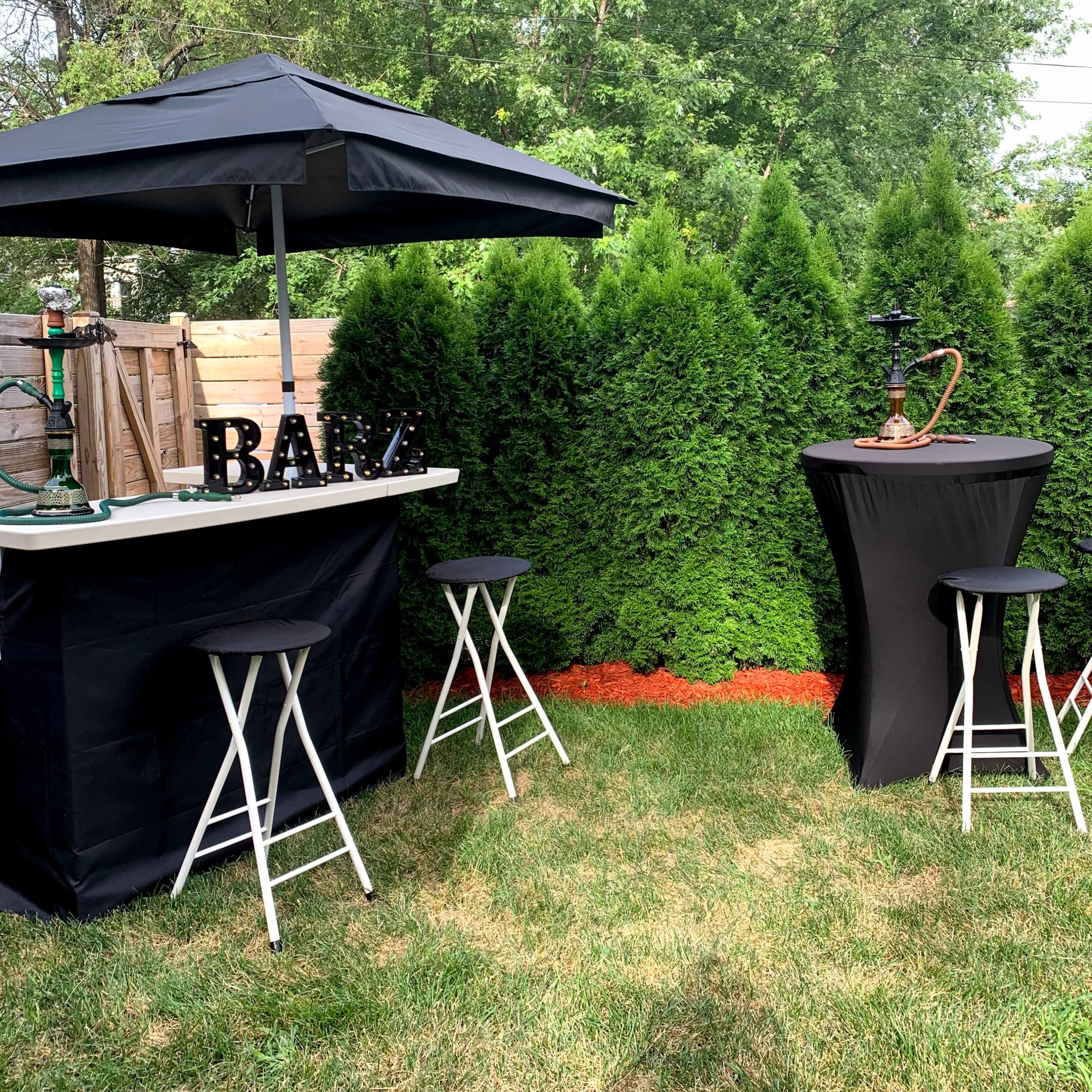 A backyard bar setup features a black canopy, a counter with the word "BAR," two high stools, and a hookah stationed beside a hedge and flower bed.