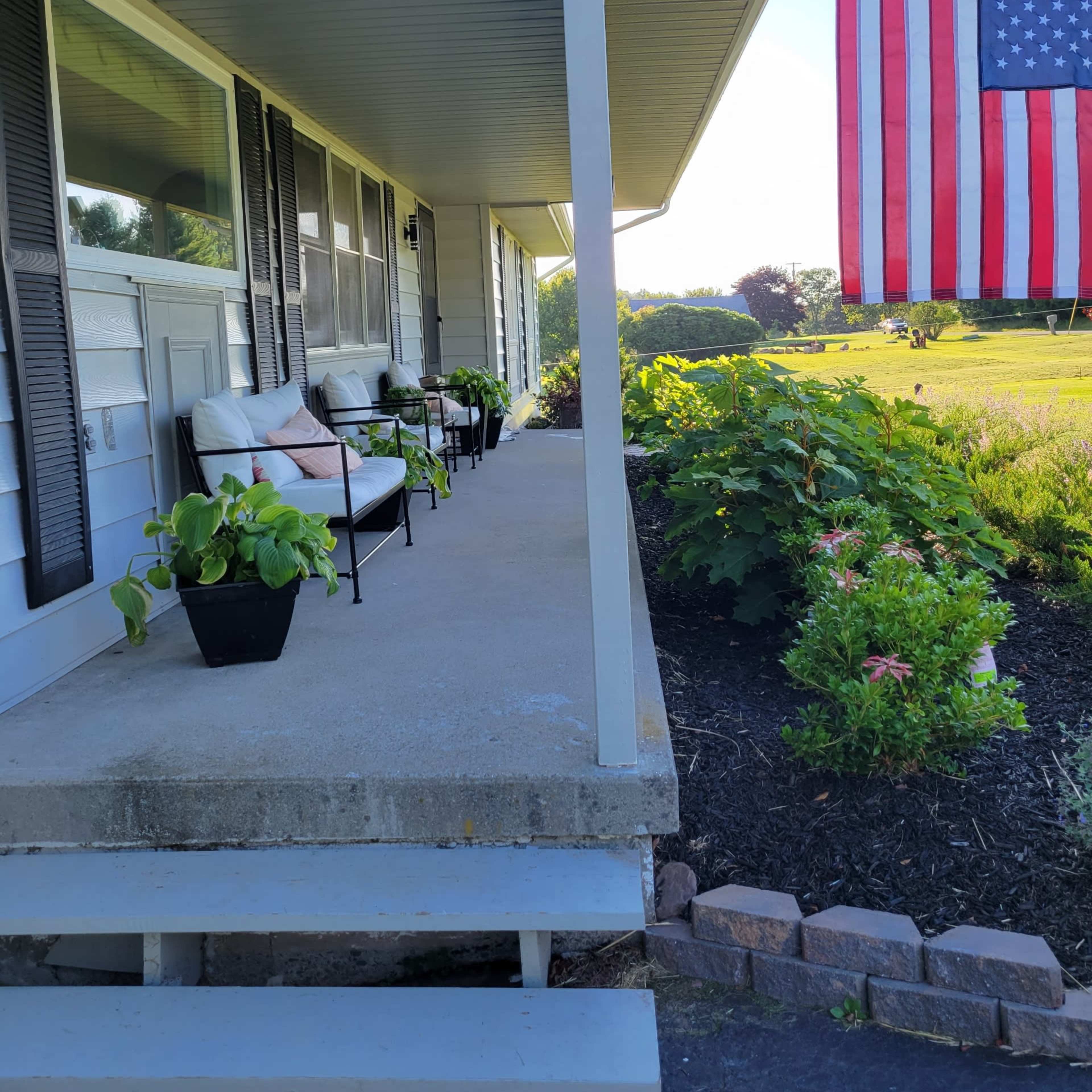 A porch with two chairs and potted plants, alongside an American flag hanging from the side of the house.