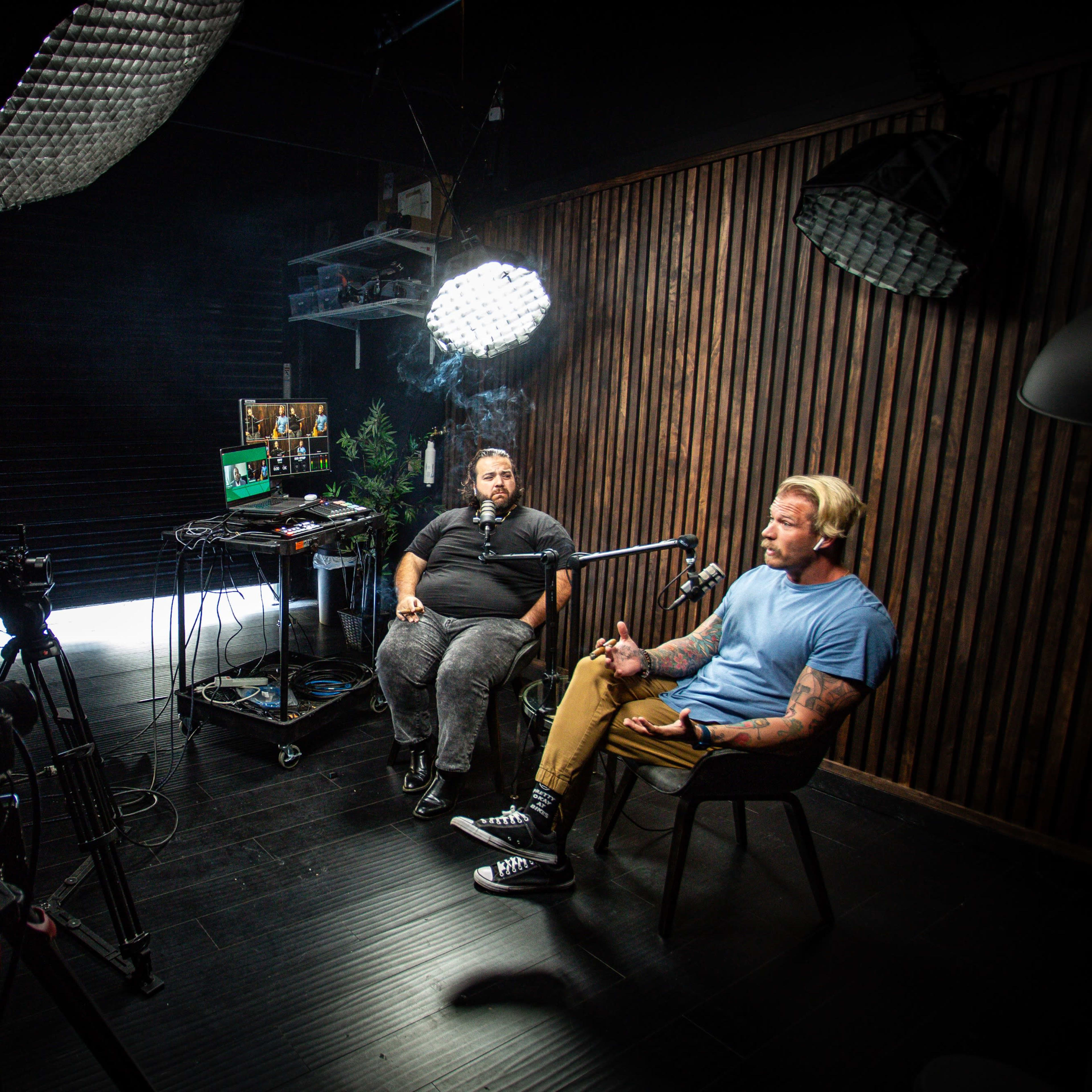 Two men are seated in a dimly lit studio, engaged in a conversation while being recorded by cameras and illuminated by softbox lights.