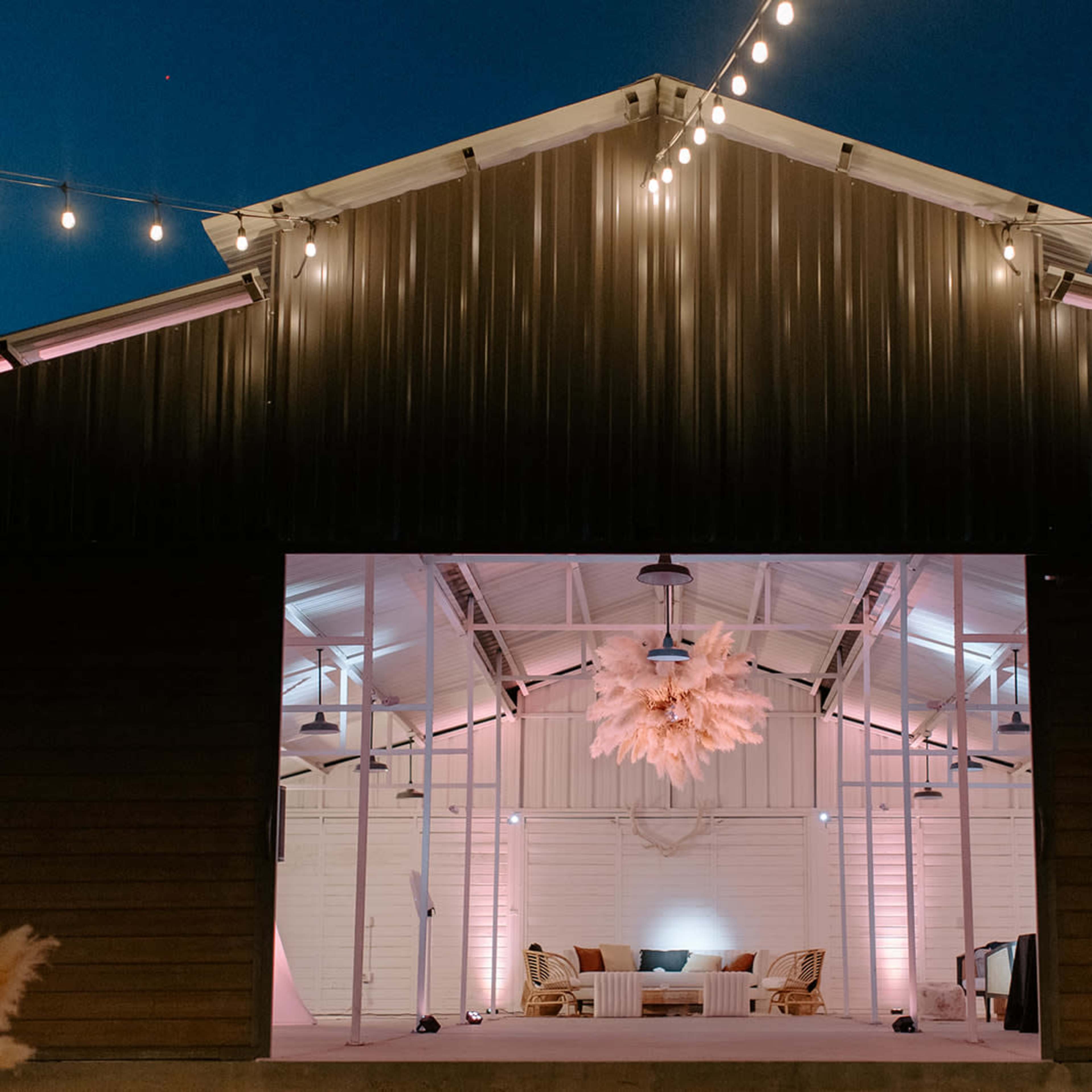 A barn with a lit outdoor seating area, featuring string lights and a large decorative chandelier, illuminated against a twilight sky.