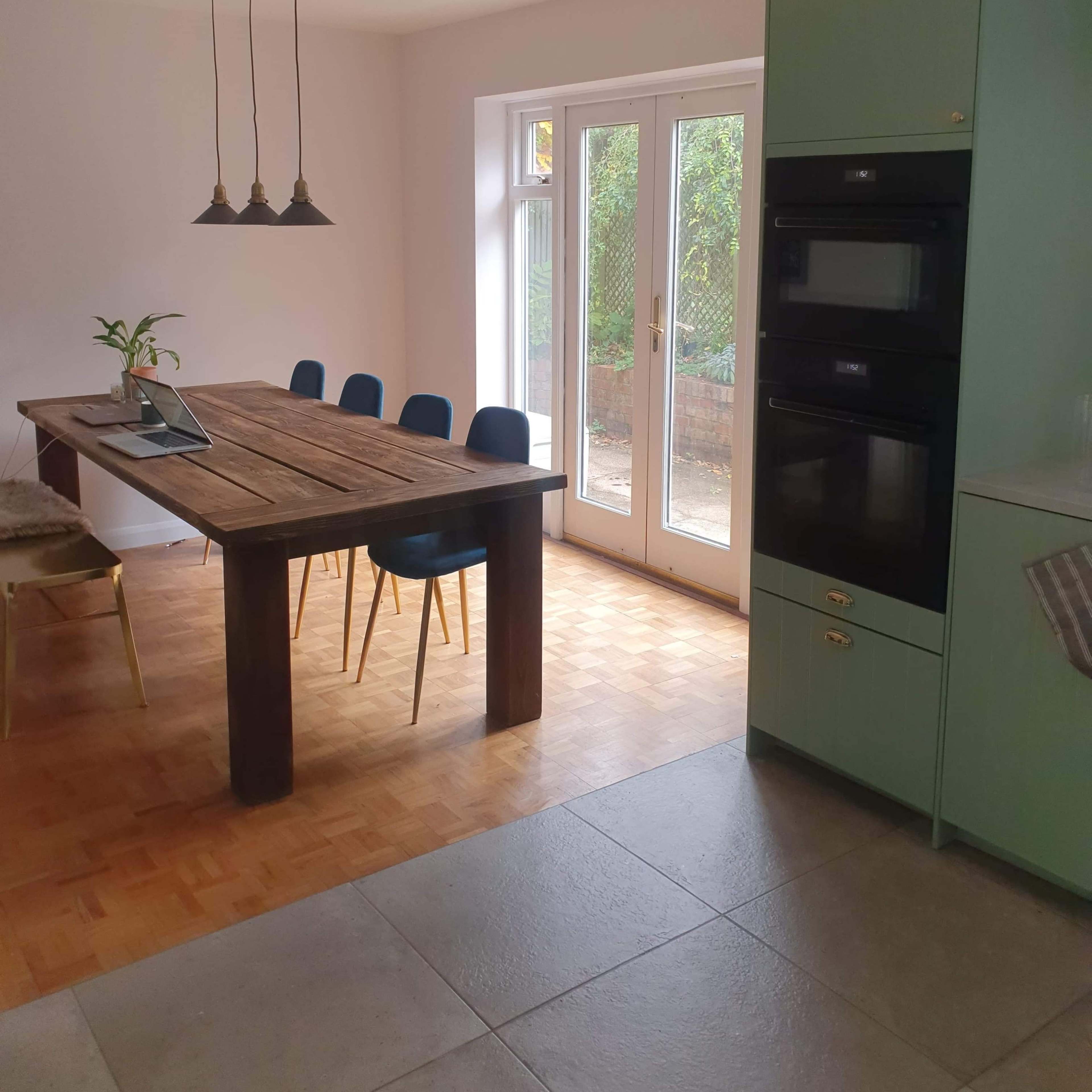 A modern kitchen with a wooden dining table, four blue chairs, and two built-in ovens adjacent to a sliding glass door.