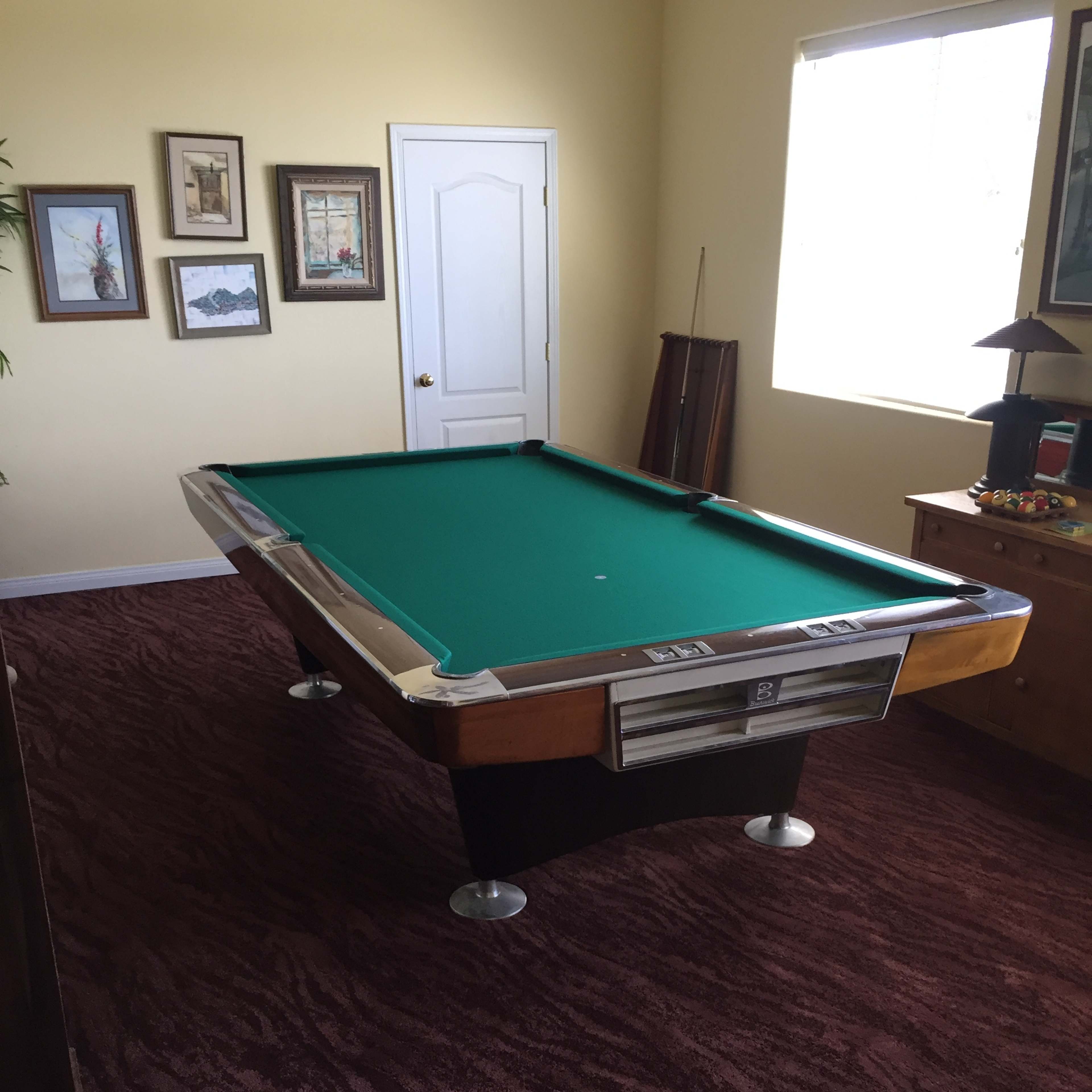 A pool table in a room with yellow walls, framed pictures, and a window providing natural light.