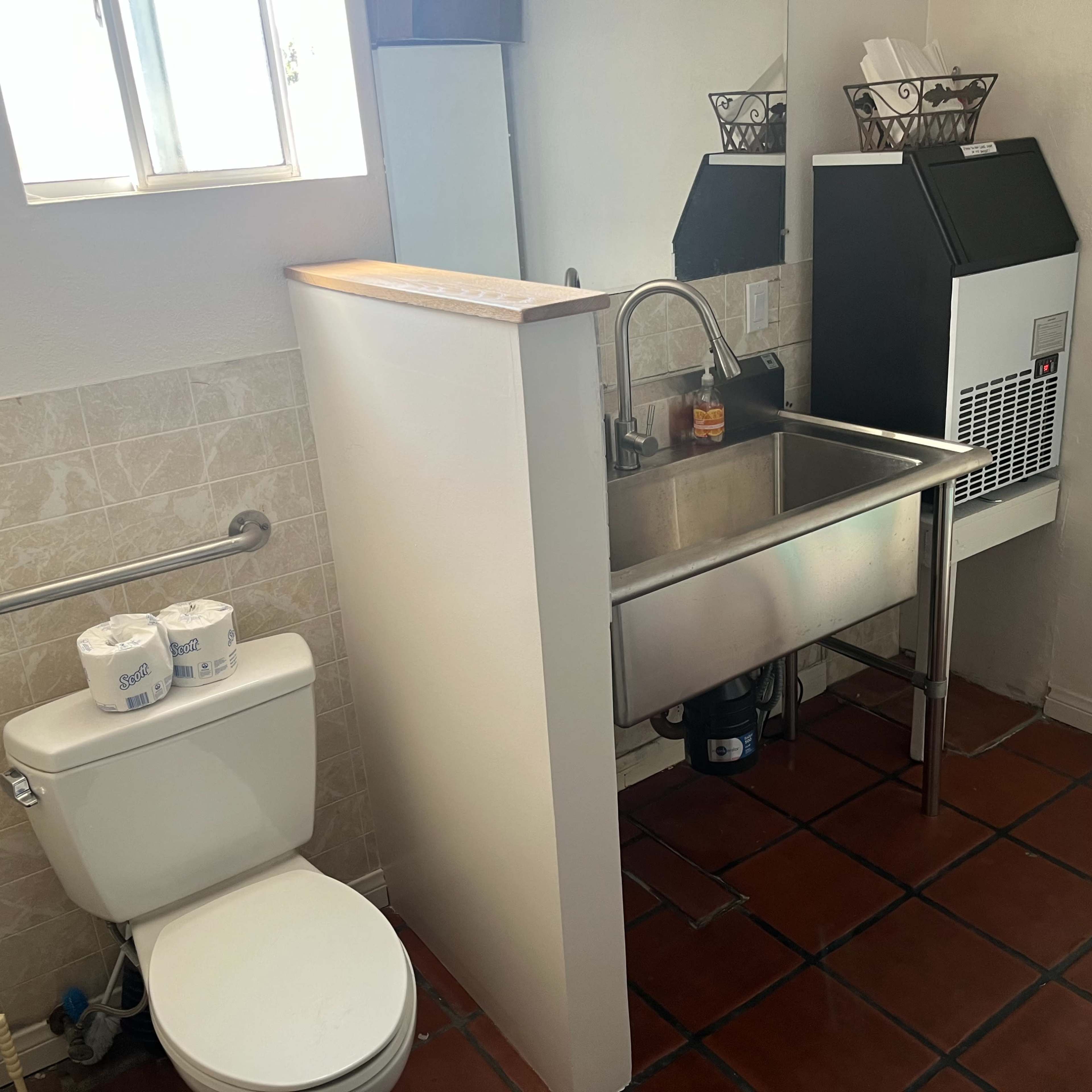 A compact bathroom with a toilet, a stainless steel sink, and a commercial ice maker on the wall opposite the window.