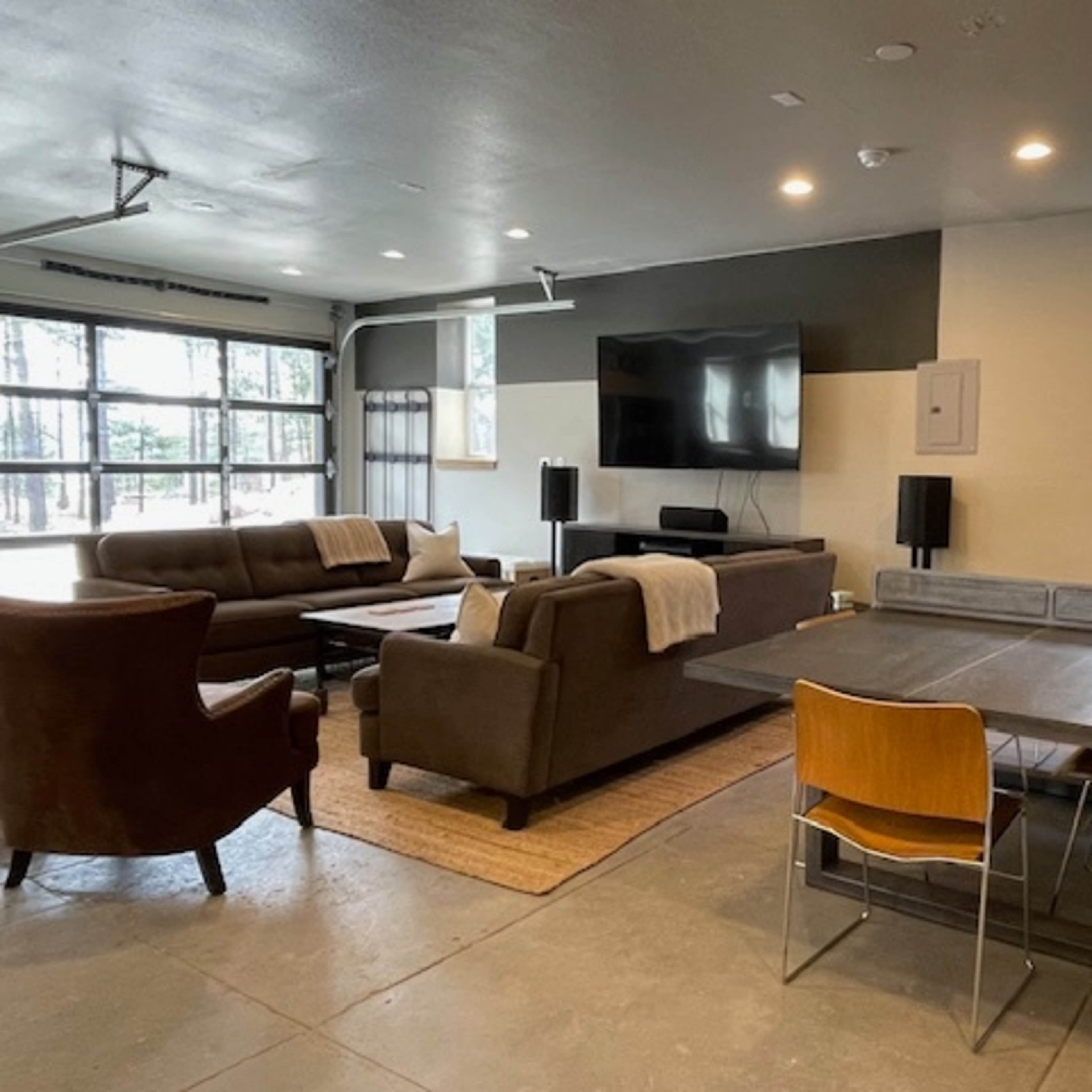 A modern living space features a sectional sofa, armchair, large TV, and dining table with chairs against a backdrop of large windows and minimalistic decor.