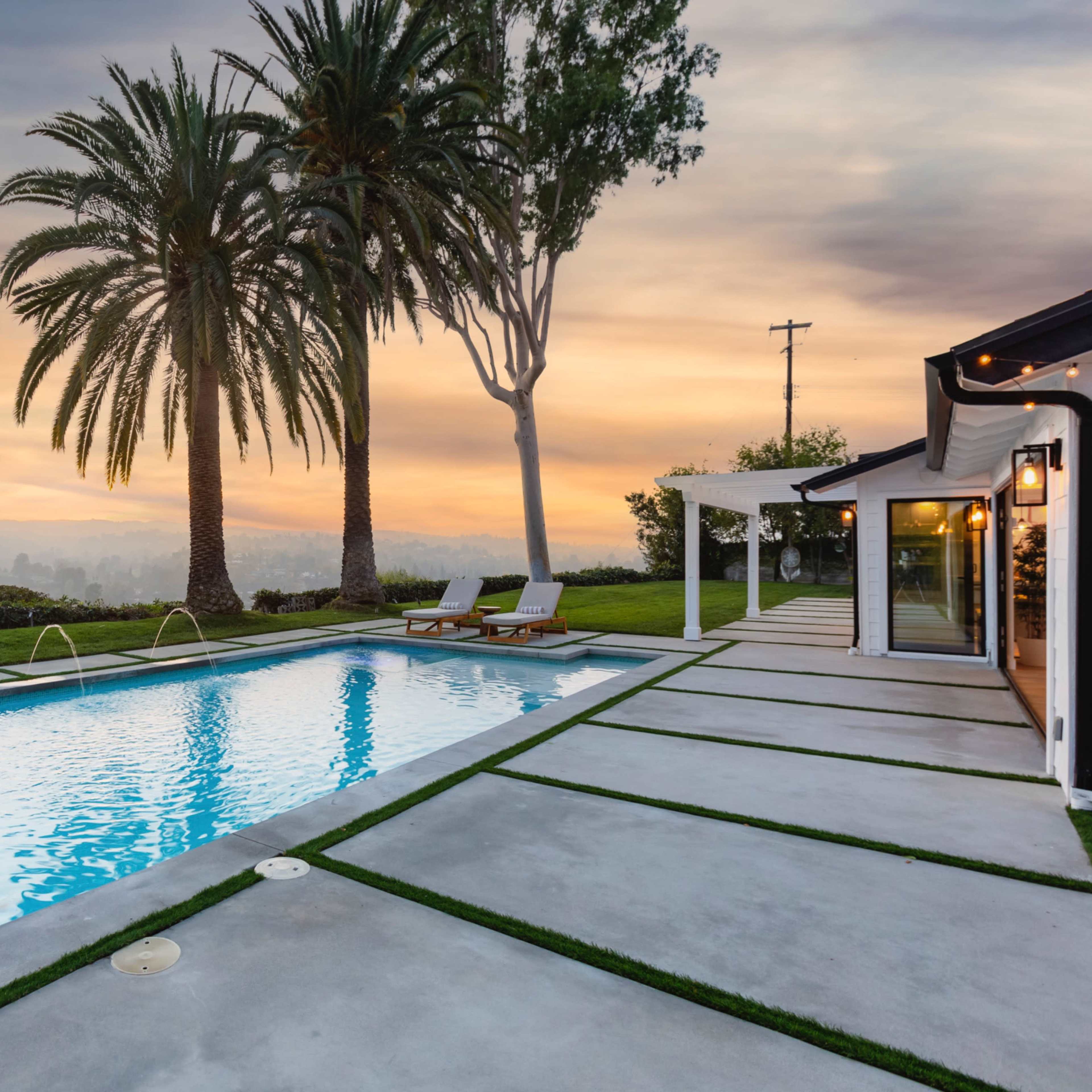 A swimming pool surrounded by palm trees and a concrete deck overlooks a scenic sunset view.