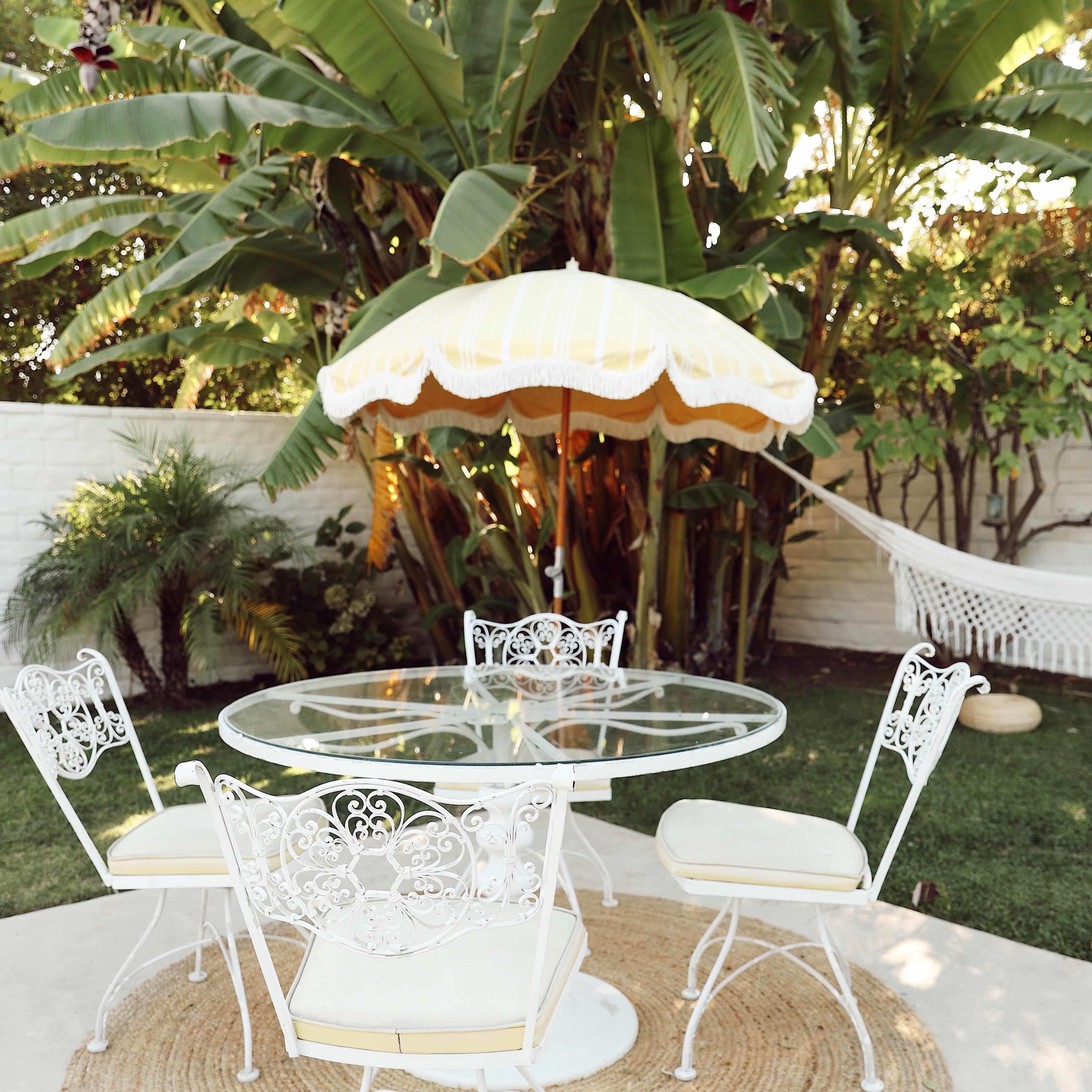 A round glass table with four white metal chairs is set under a yellow-striped umbrella in a backyard surrounded by lush greenery, including banana plants and a hammock.