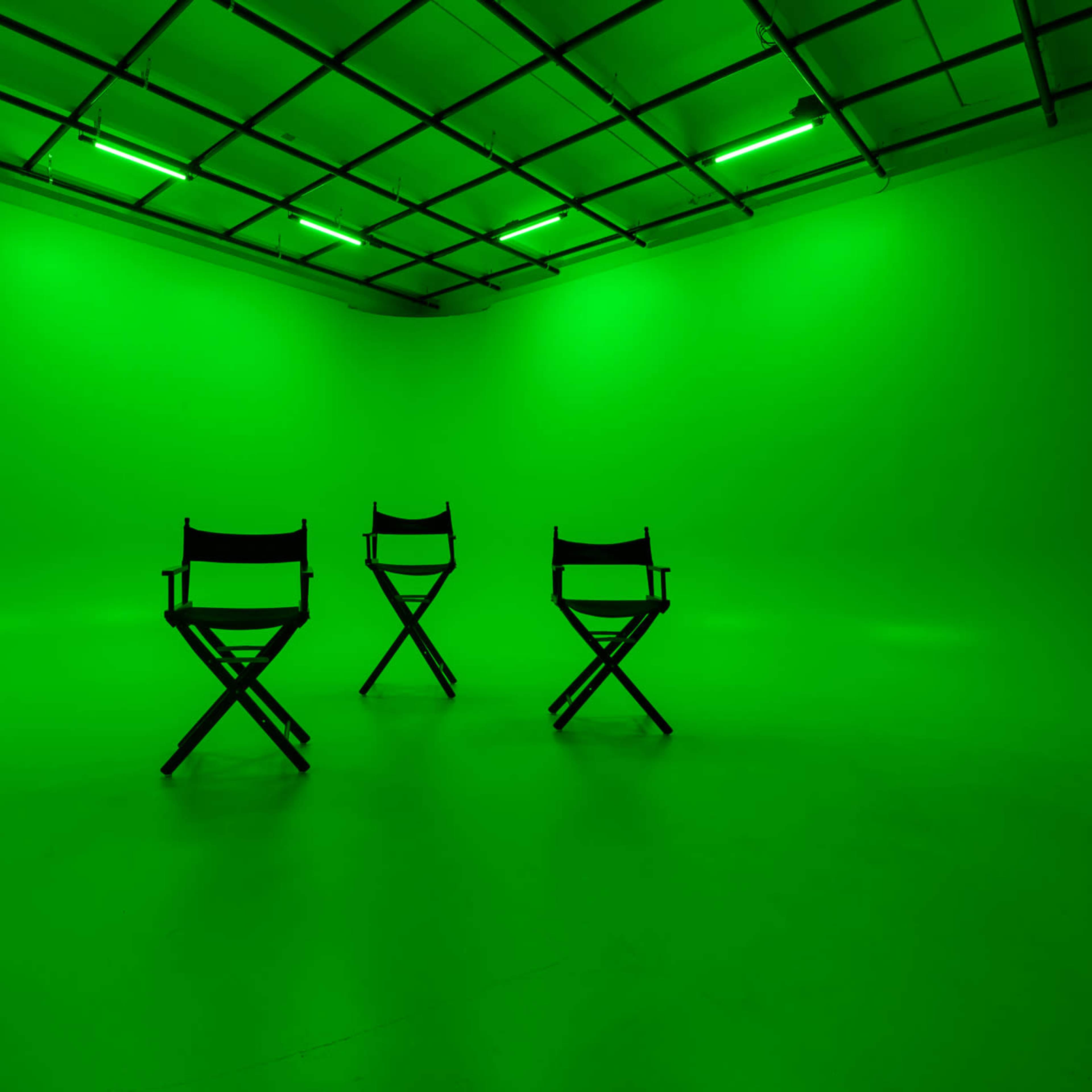 Three empty director's chairs are arranged in a brightly lit green studio space.