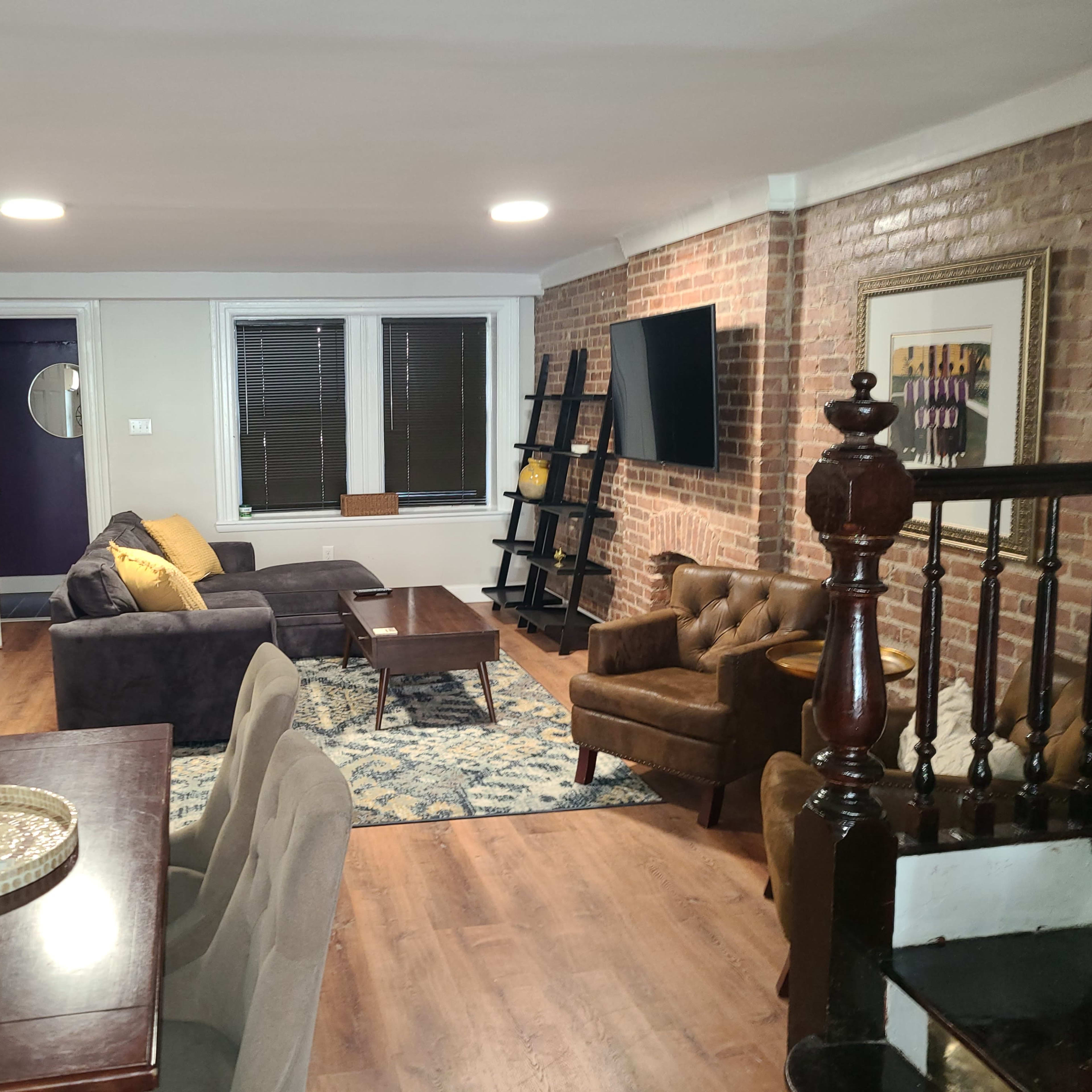 A modern living room with a brick wall, a sofa, armchair, coffee table, and dining area, illuminated by overhead lights.