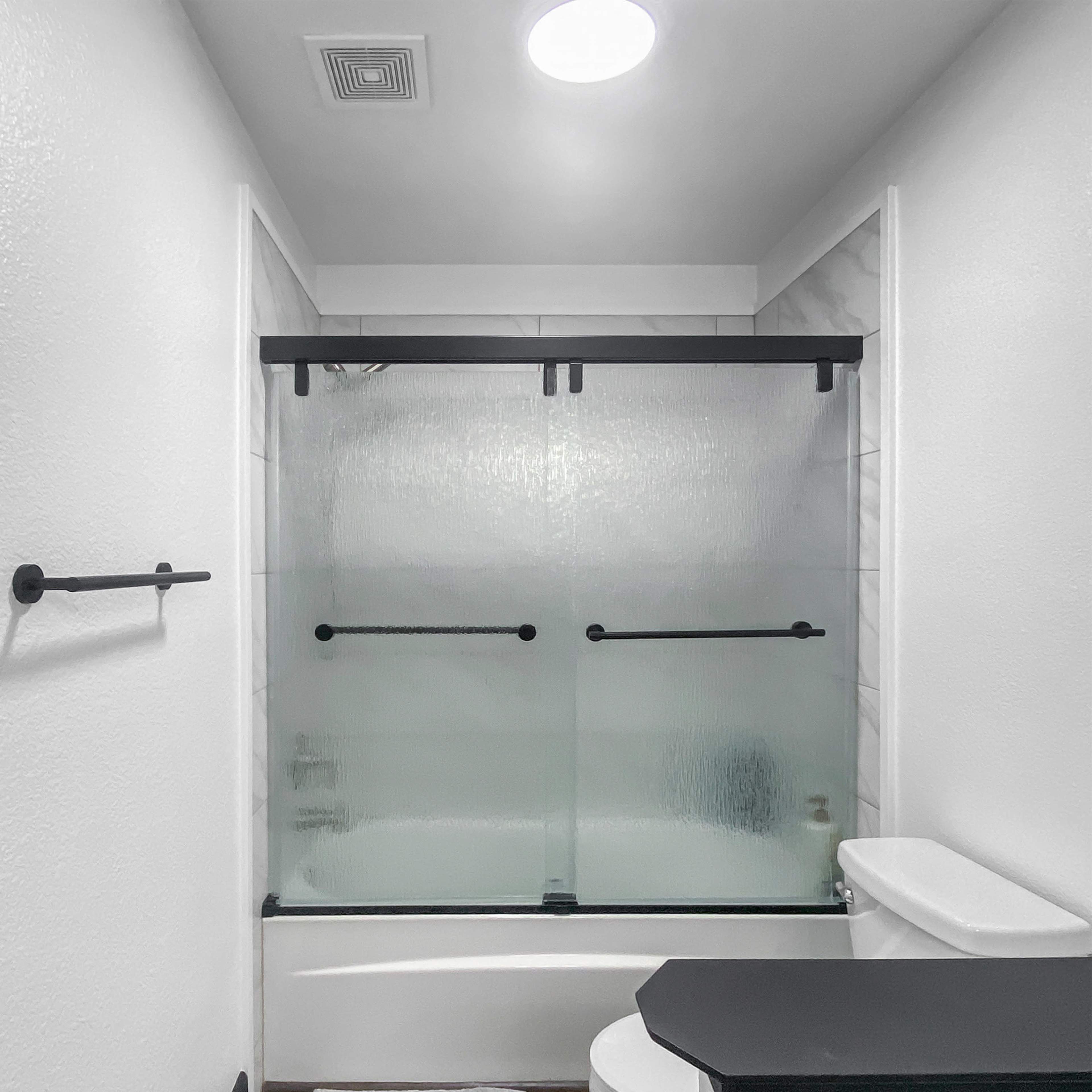 A modern bathroom features a sliding glass shower door, a white toilet, and a black countertop beside a tub.