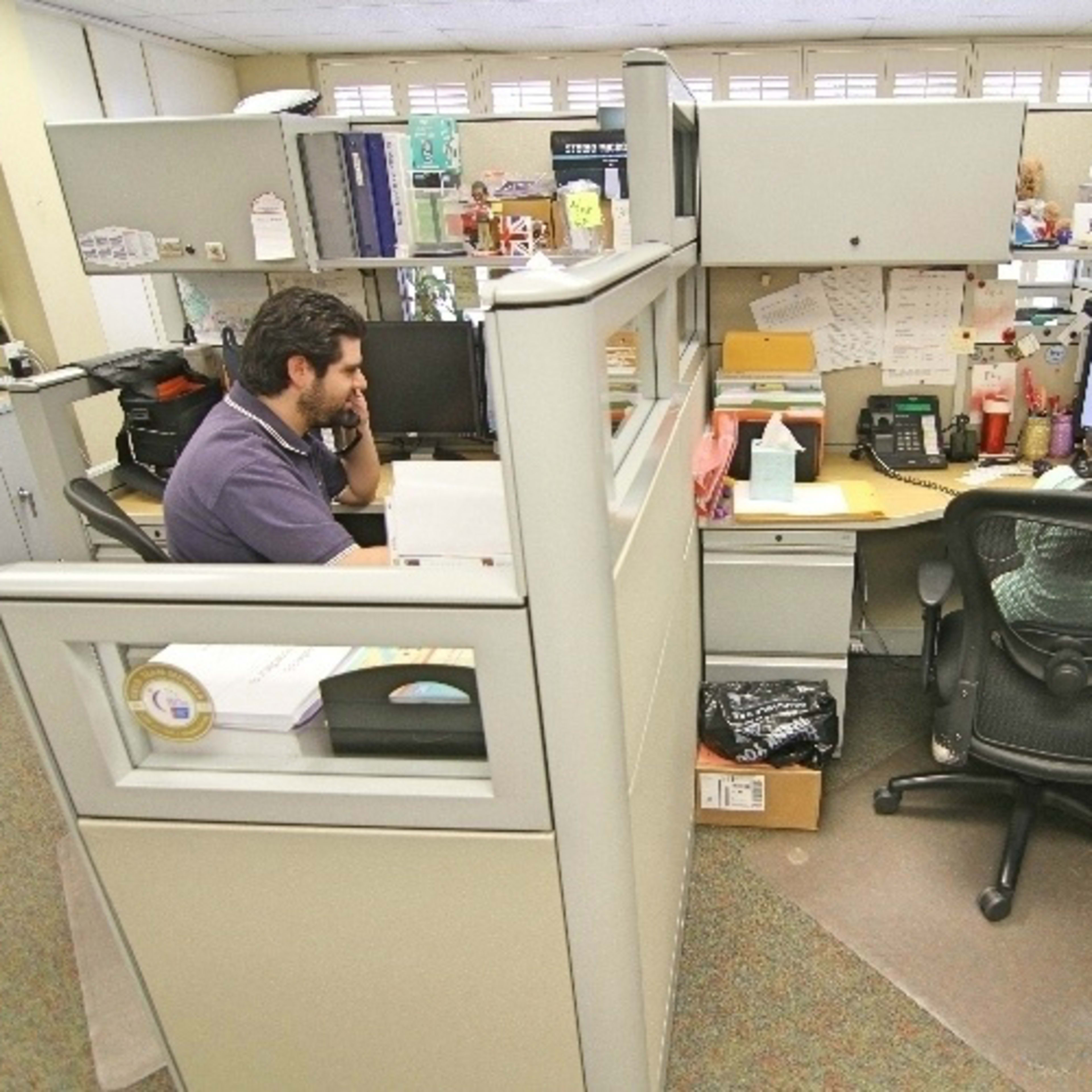 Two office workers are seated at adjacent cubicles, one talking on a phone and the other focused on a computer screen.